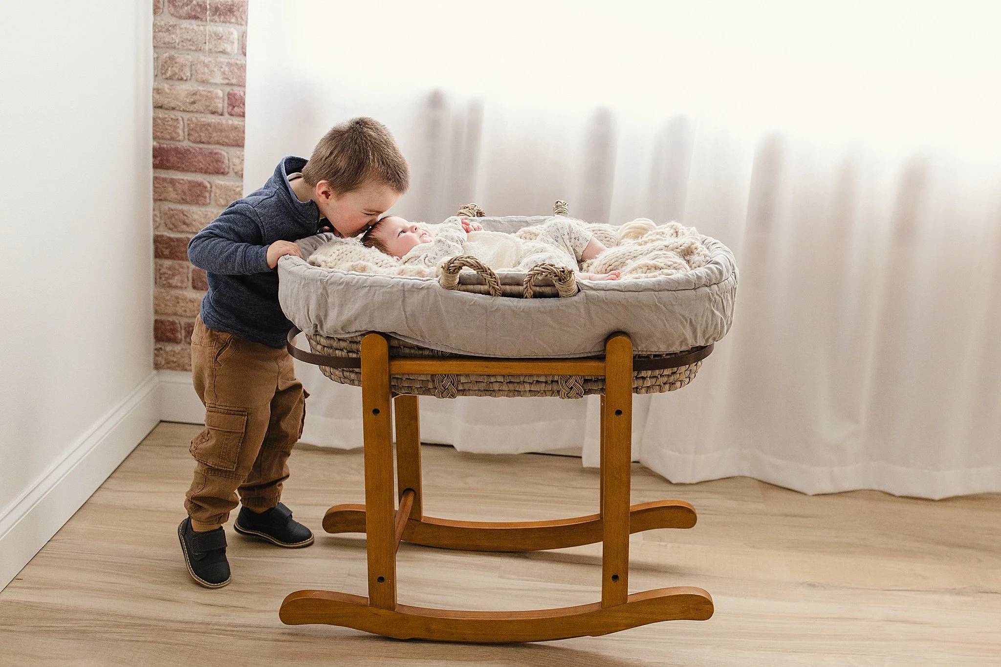 A young boy leaning over a baby lying in a wicker bassinet, in a room with light-colored walls, wooden flooring, and a partial brick wall in the background with a Naperville newborn photographer.