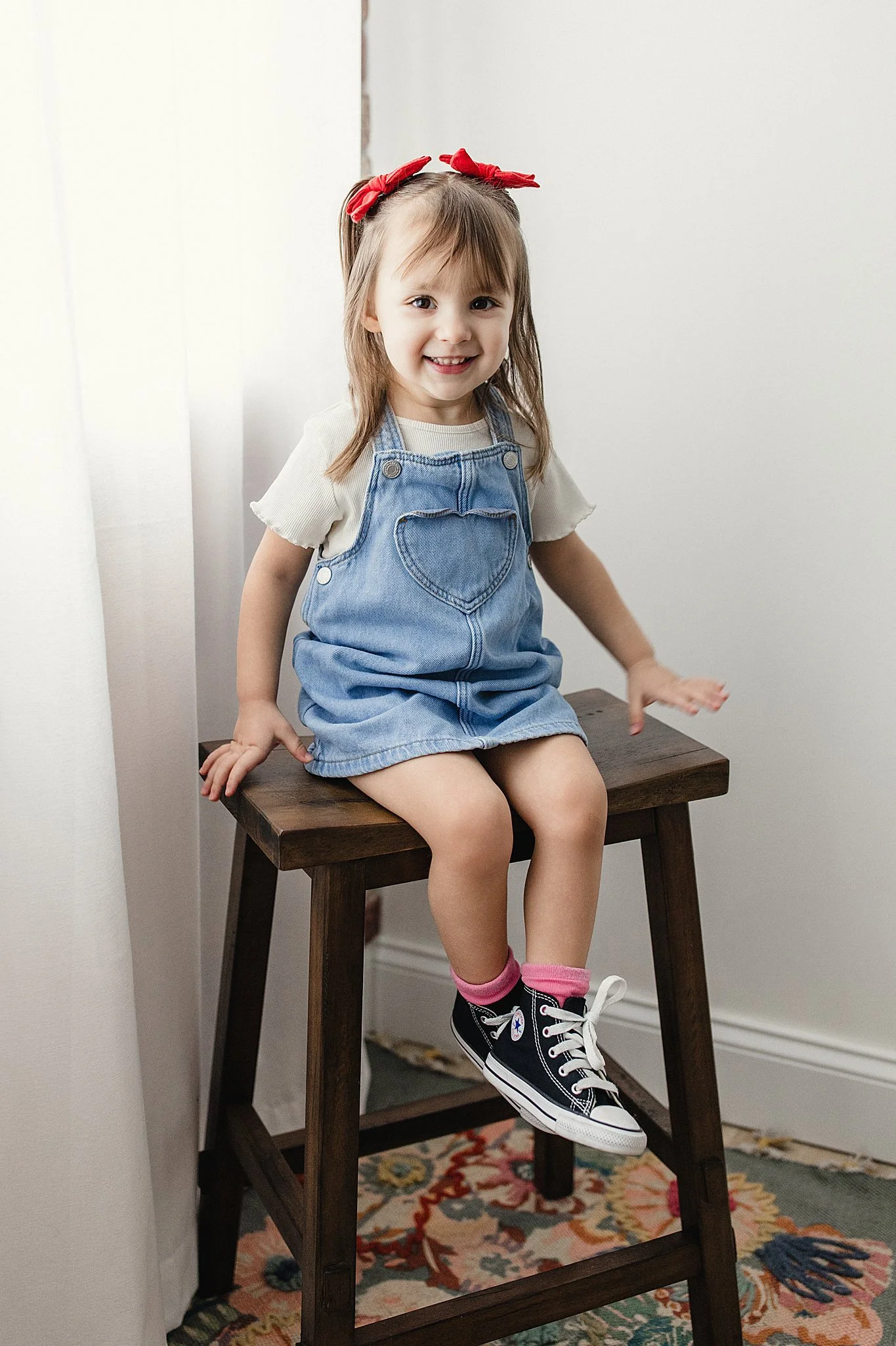 A young girl with a red bow in her hair is sitting on a wooden stool, smiling, wearing a beige shirt, a denim dress with a heart-shaped pocket, pink socks, and black sneakers, in a room with white walls and a colorful rug for kids photography near me
