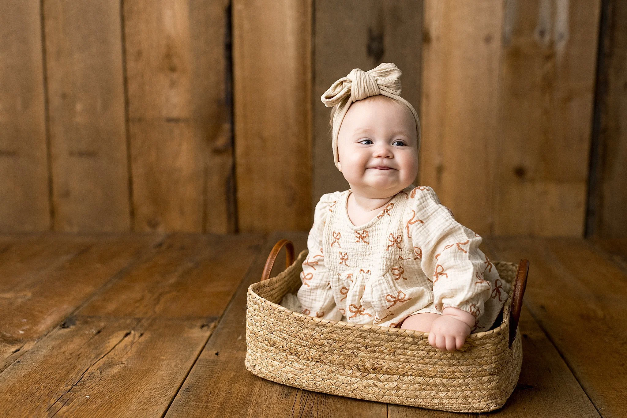 A baby girl with a beige headband tied in a bow, sitting in a woven basket on a wooden floor with a wooden wall background. She's wearing a cream dress and has a slight smile, sticking her tongue out slightly for baby photos Naperville.