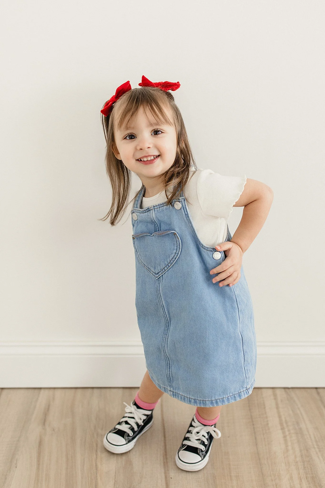 A young girl with light brown hair, red bow, and a white shirt, wearing a denim dress with a heart-shaped pocket, black and white sneakers, and pink socks during kids photography in Naperville, IL.