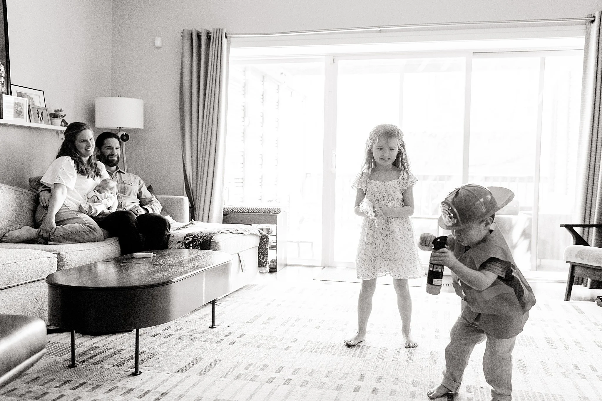 A family sitting on a couch in a living room watching children playing. One child is holding a toy gun, and another child is standing and smiling while holding a stuffed toy. The room has a large window with curtains, and there are decor items on a s