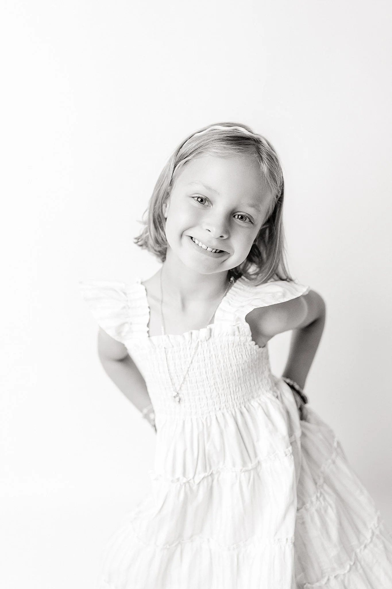 A young girl with shoulder-length hair and a big smile, wearing a light-colored dress with ruffled details, standing against a plain white background for a personality mini session in Naperville.