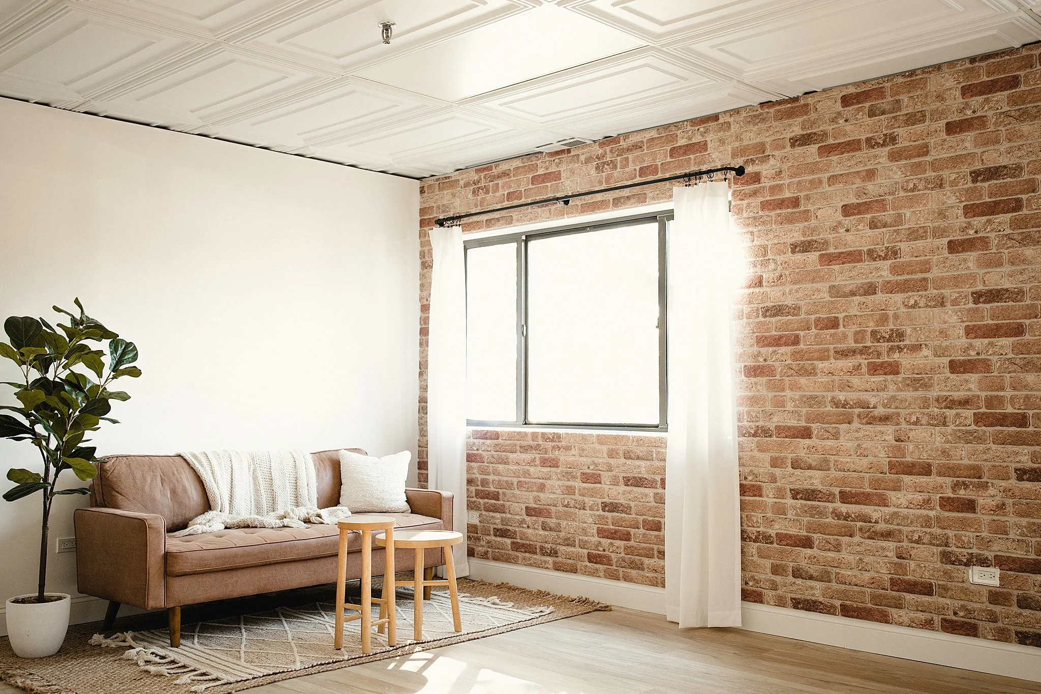 Living room with a brick wall, a brown sofa with decorative pillows, a potted plant, and wooden tables, illuminated by natural light through a window with white curtains in a photo studio near me.