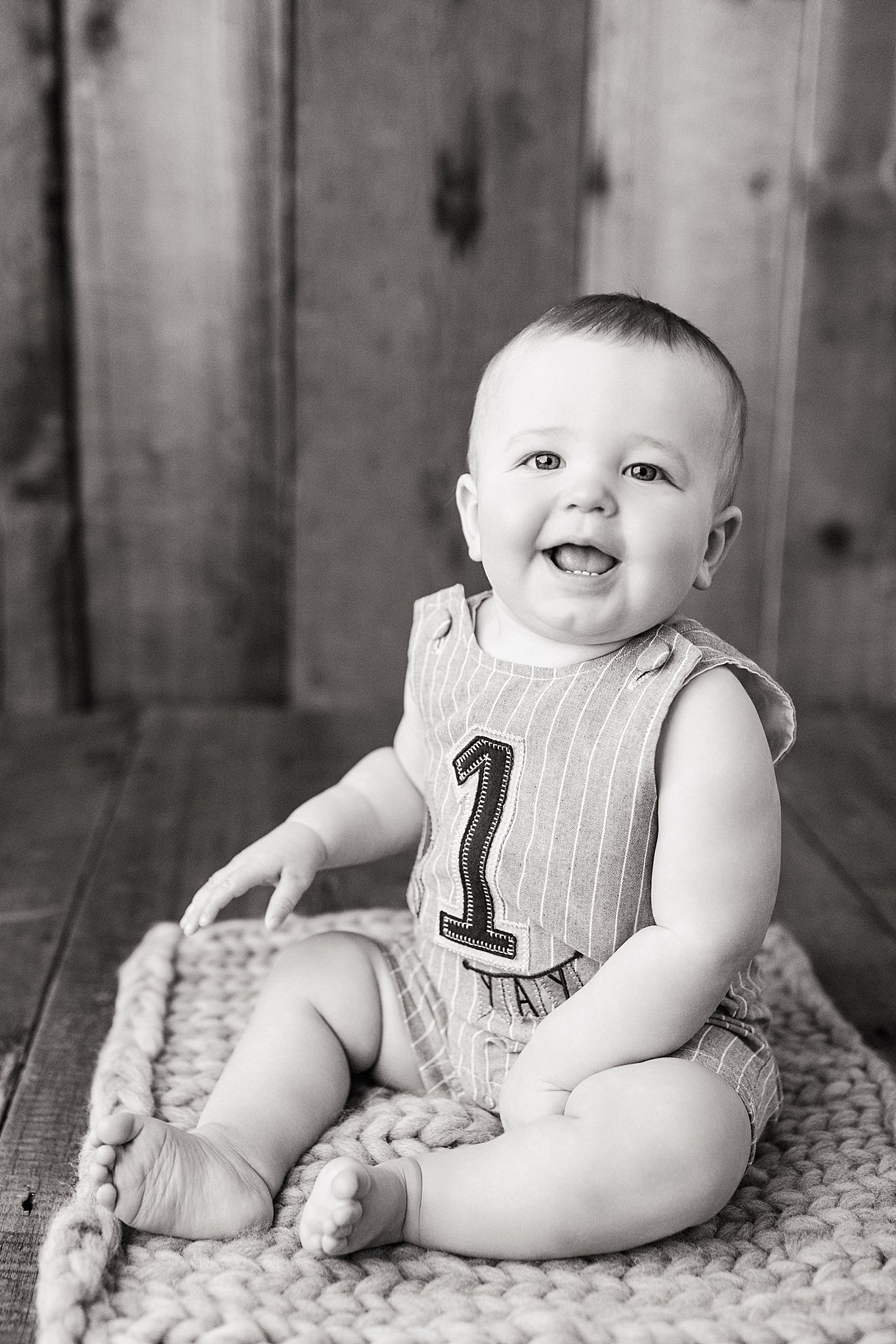 A smiling baby sitting on a textured blanket, wearing a striped outfit with the number 1 on it, in front of a wooden background during a milestone photoshoot with Ally and B Photography.
