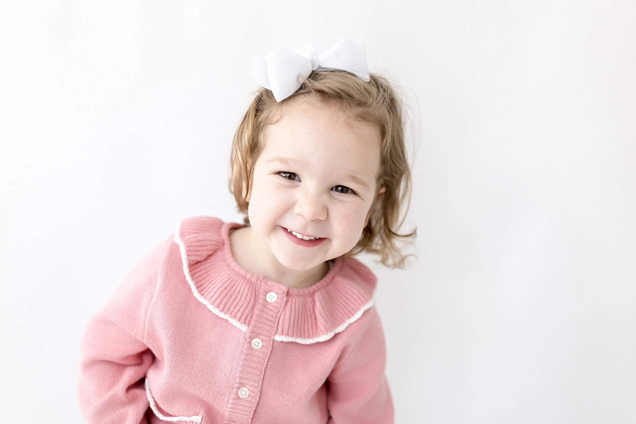 A young girl with light brown hair in loose curls, wearing a pink sweater with a ruffled white collar and white buttons, smiling at the camera against a plain white background, with a white bow with the best kids photographer in Naperville.