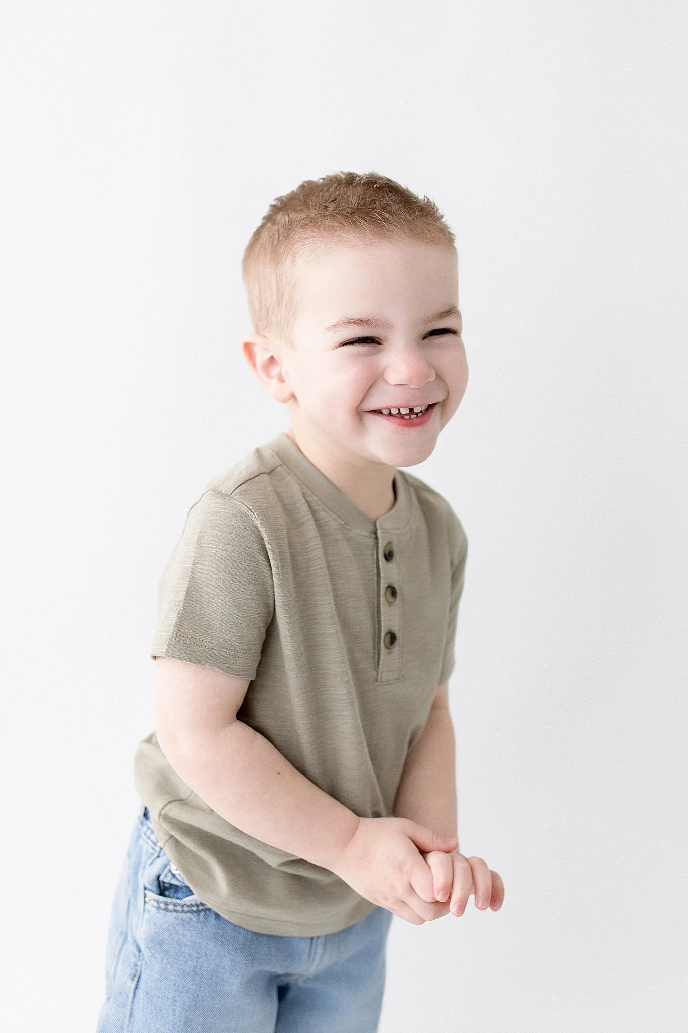 A young boy smiling and laughing with hands clasped in front of him, wearing a beige shirt and blue jeans, with a plain white background with the best kids photographer Naperville.