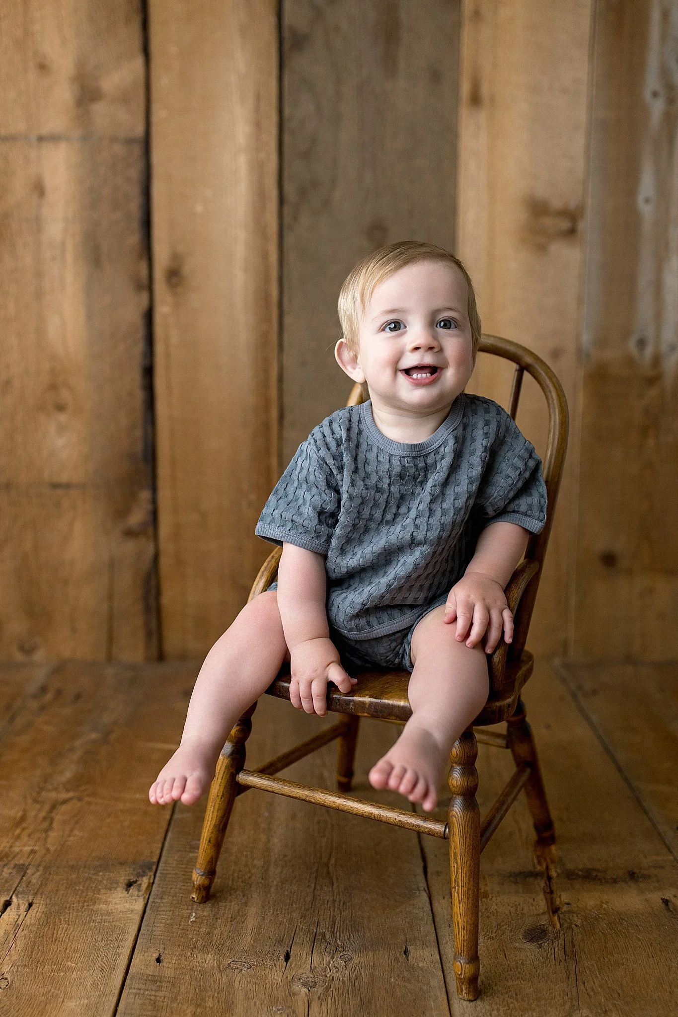 A young smiling child with light brown hair and blue eyes sitting on a small wooden rocking chair in front of a wooden wall, wearing a gray t-shirt during milestone photography Naperville.