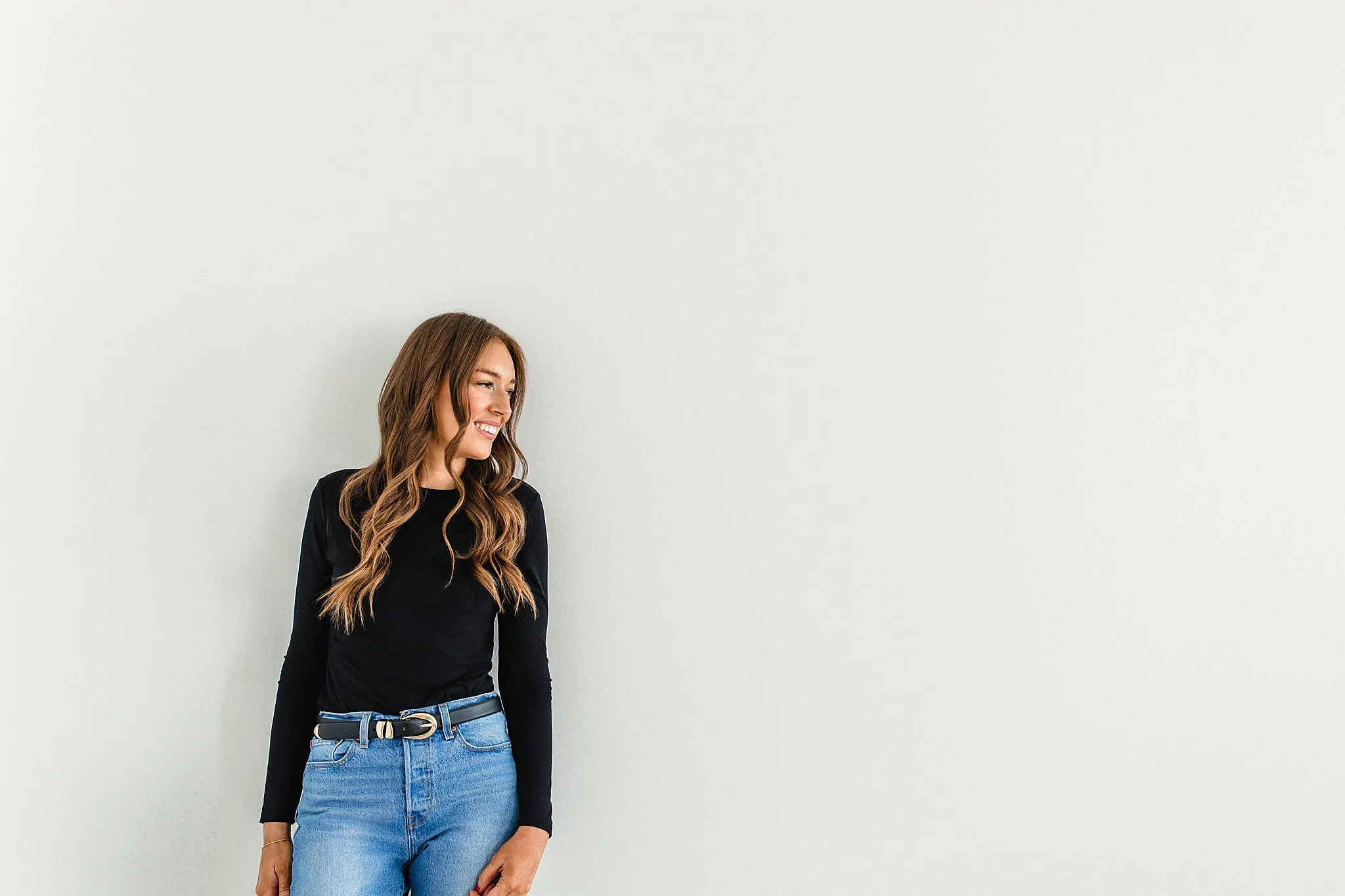A woman with long brown hair, wearing a black long-sleeve top and blue jeans, standing against a plain white wall, smiling and looking to her right for Naperville branding photos.