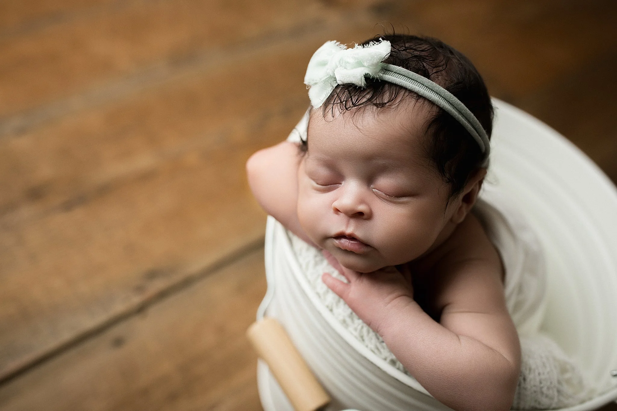 A sleeping baby with curly dark hair wearing a headband, resting on a white chair with a wooden surface nearby for baby photos near me.
