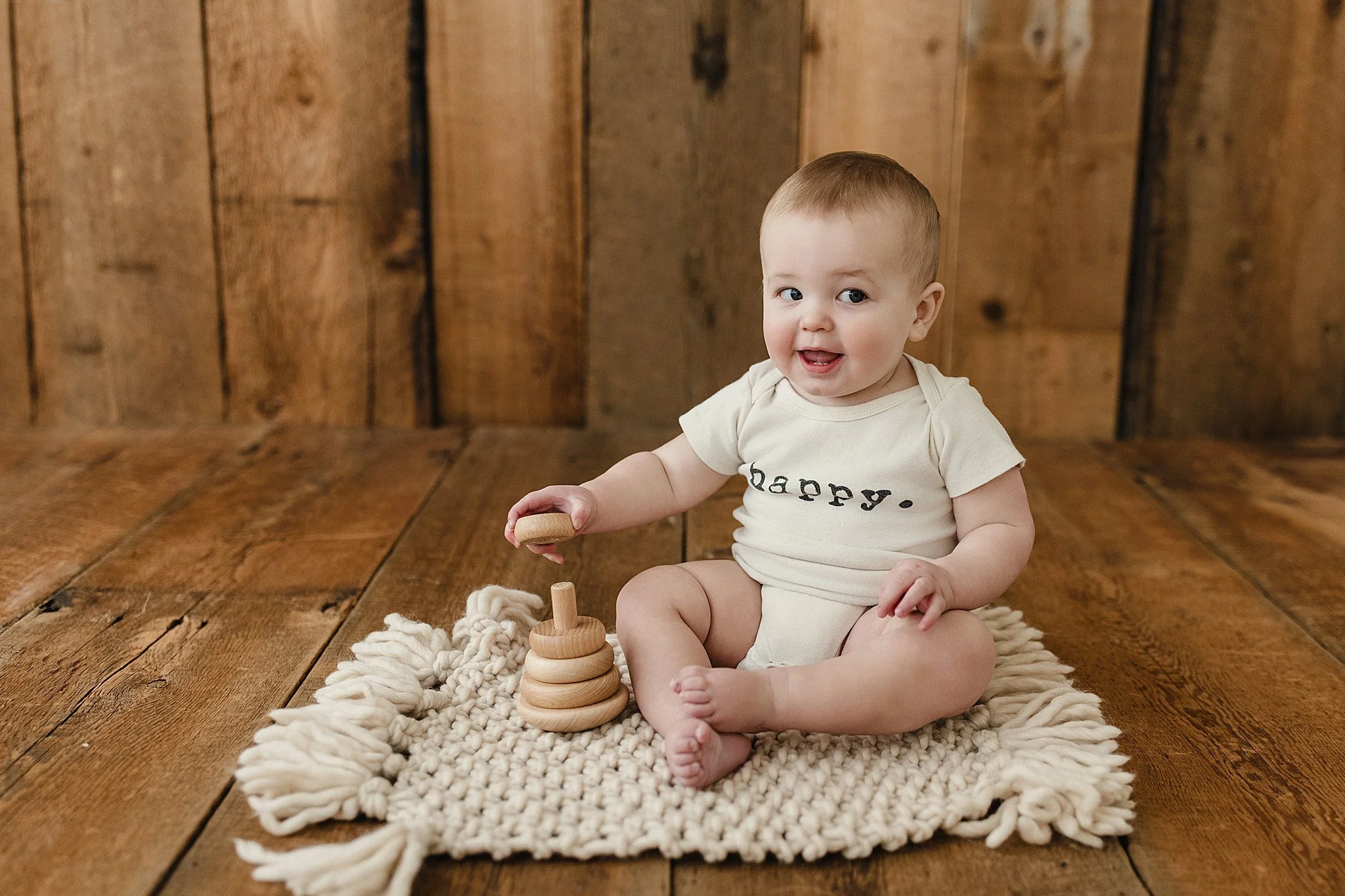 A smiling toddler with short light hair sitting on a cream-colored knitted rug on a wooden floor, playing with a wooden stacking toy, wearing a cream-colored onesie with the word 'happy.' printed on it, against a wood backdrop during one year photos.