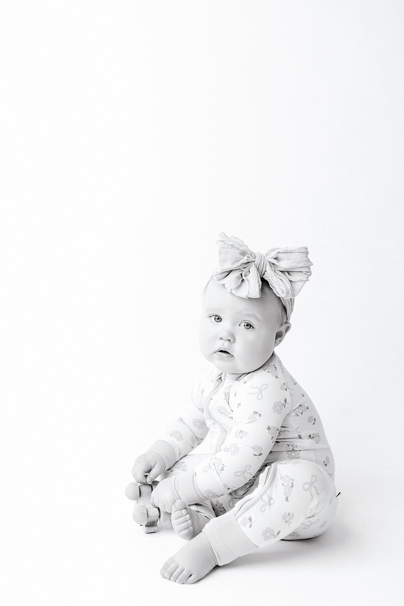 A baby sitting on the floor, wearing a long-sleeved onesie with a floral pattern and leggings. The baby has a bow headband and is holding a small toy. The background is plain white with Ally and B Photography baby photos.