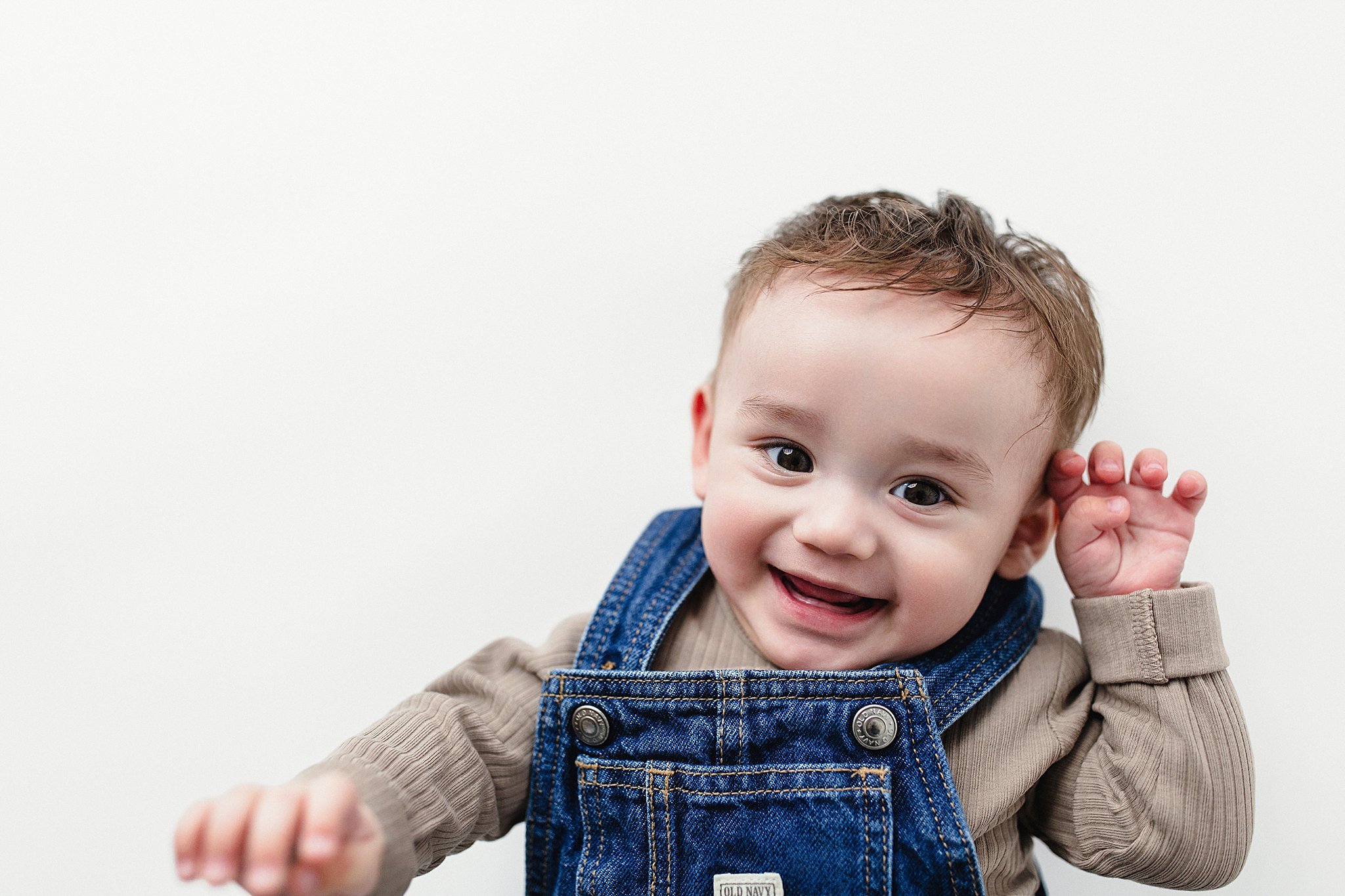 Happiness of a young boy with brown hair, smiling, wearing a blue denim overall with a beige long-sleeve shirt, against a plain white background.