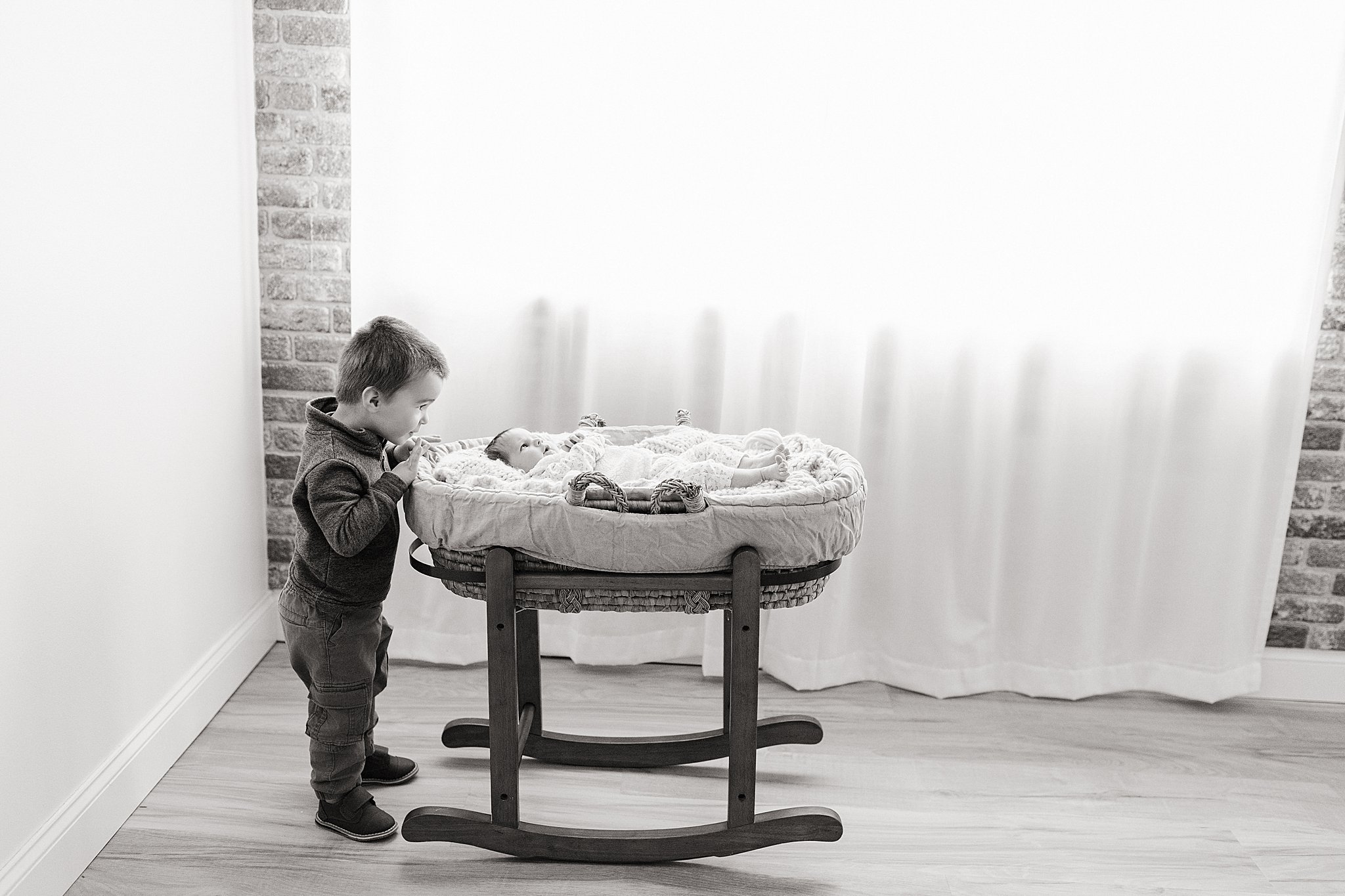 A young boy looking at a newborn baby lying in a bassinet in a room with white walls, a brick wall, and a window with curtains during family photos in naperville.