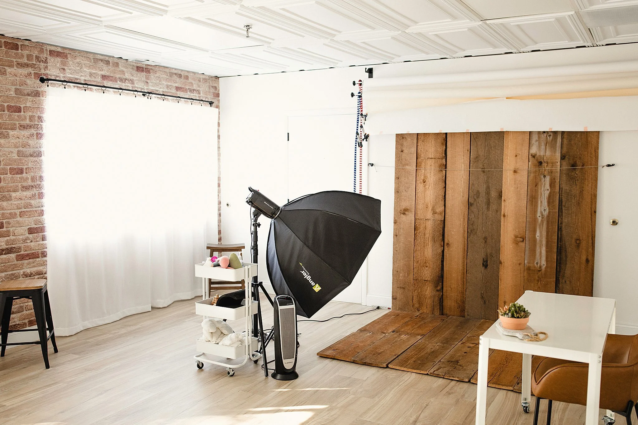 Photo of a photography studio with a brick wall, a window with white curtains, a photography backdrop, a large softbox light, and a cart with photography equipment, a small table with a plant, and a chair in photo studios near Naperville.