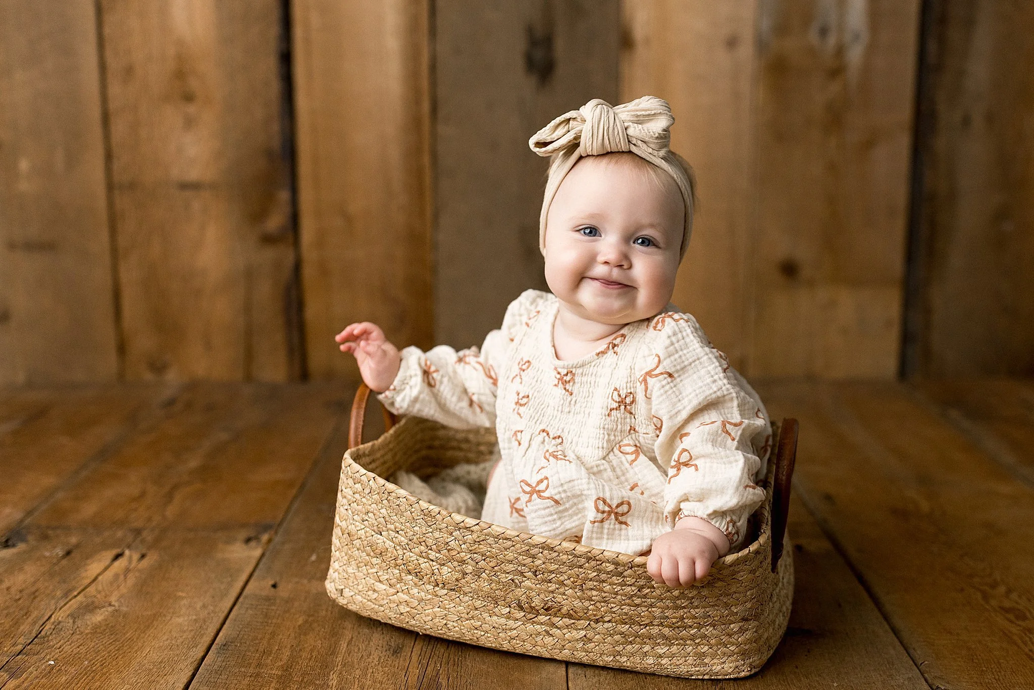 A baby girl with blue eyes and a beige headwrap with a bow, sitting in a woven basket on a wooden floor, smiling at the camera during baby photos with Ally and B Photography.