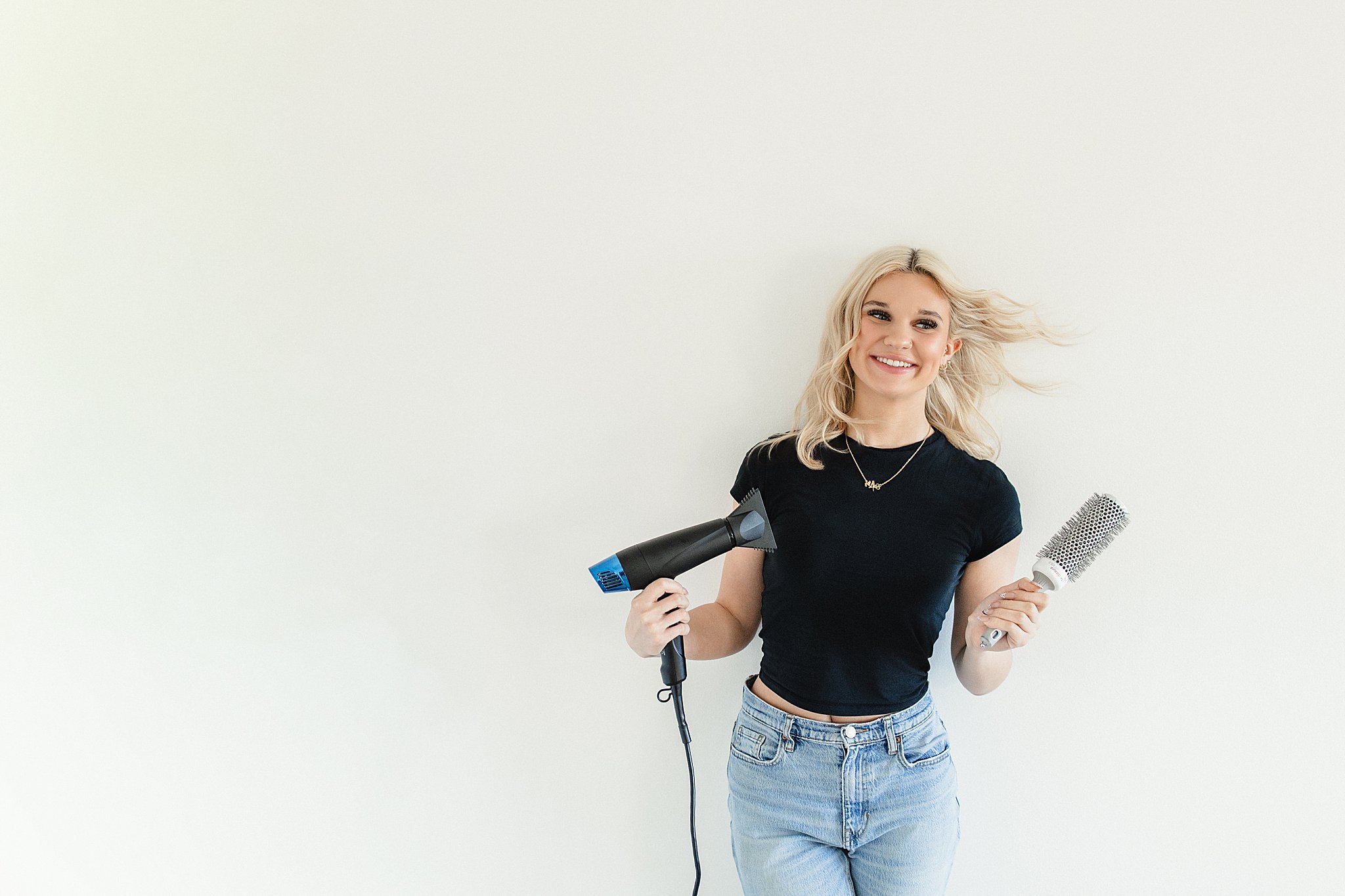 Young woman with blonde hair holding a blow dryer and a round hairbrush, smiling, standing against a plain white background for a hairstyle branding sesison.