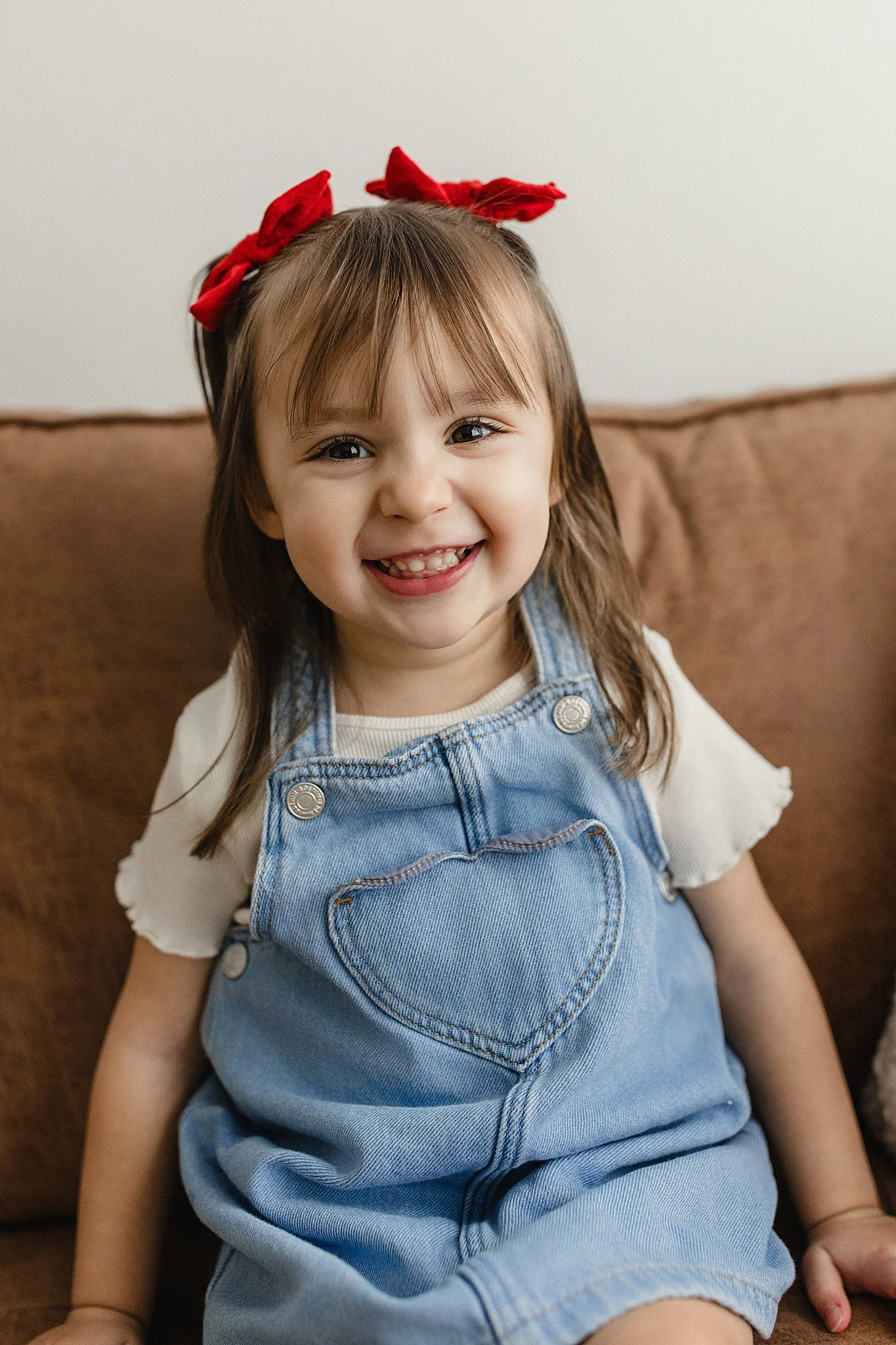 A young girl with brown hair and a red bow, smiling and sitting on a brown couch, wearing a white shirt and denim overalls for a kids model session in Naperville.