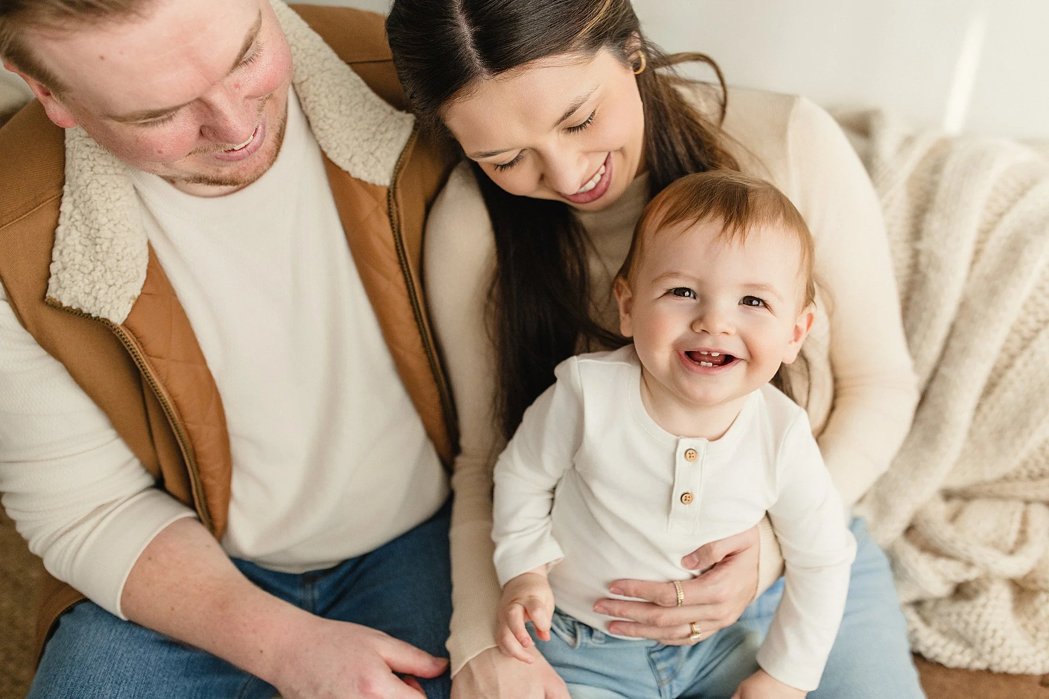 A happy family sitting on a couch, with a smiling toddler in front, a woman and a man behind her, all smiling and enjoying a moment together during a lifestyle photoshoot in Naperville, IL.