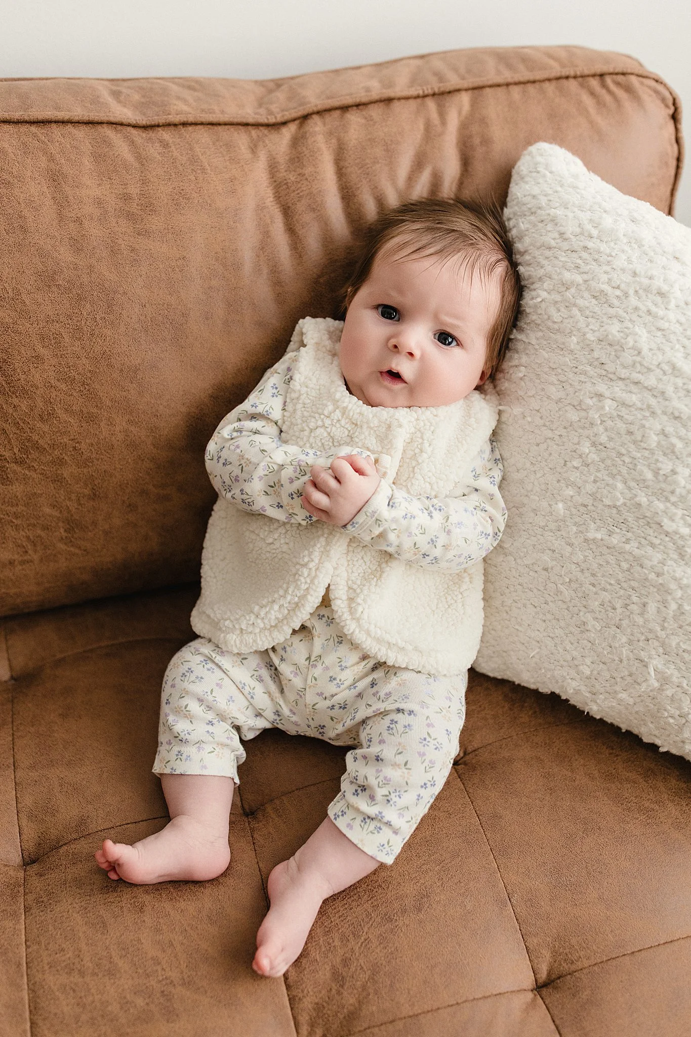 A baby sitting on a brown couch with a cream-colored fuzzy pillow, wearing a light-colored outfit with a floral pattern and a cream fleece vest, looking at the camera with a curious expression with a baby photographer near me.