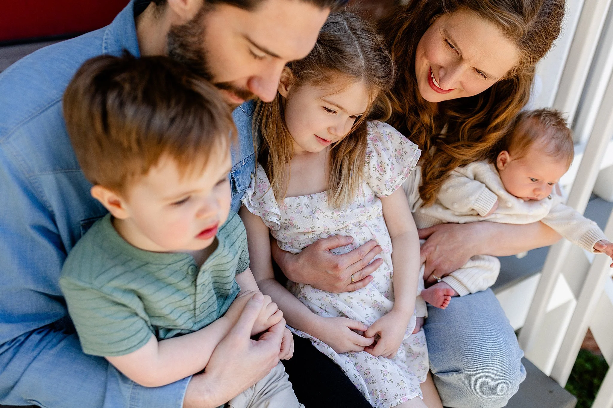 A family of six sitting closely together on a white outdoor bench during daytime, with the mother holding a baby and all family members looking downward, sharing a moment of closeness.