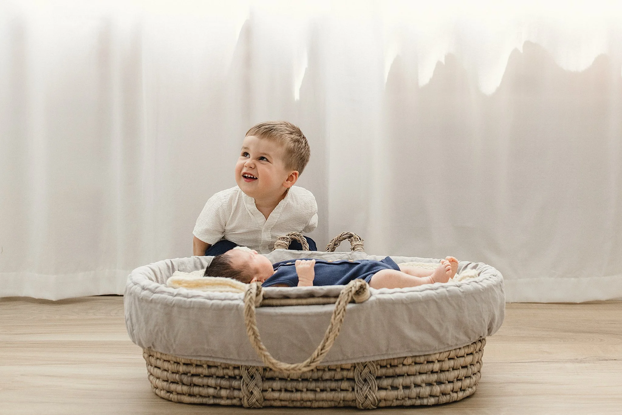 A young boy sitting next to a newborn baby on a soft blanket in a wicker basket in a bright room with white curtains with Ally and B Photography.