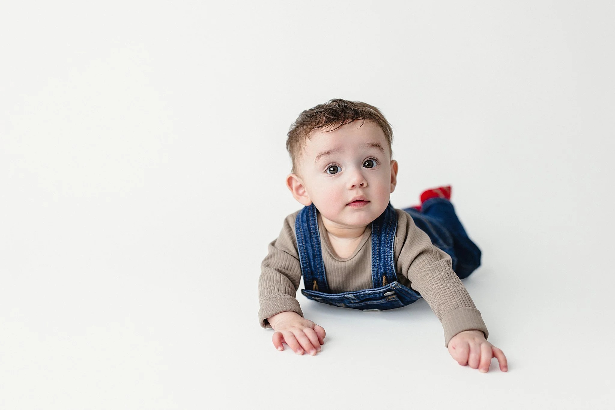 Young child with brown hair, wide eyes, and light skin, crawling on a white surface, wearing a tan long sleeve shirt, blue overalls, red socks, and dark pants, looking at the camera.