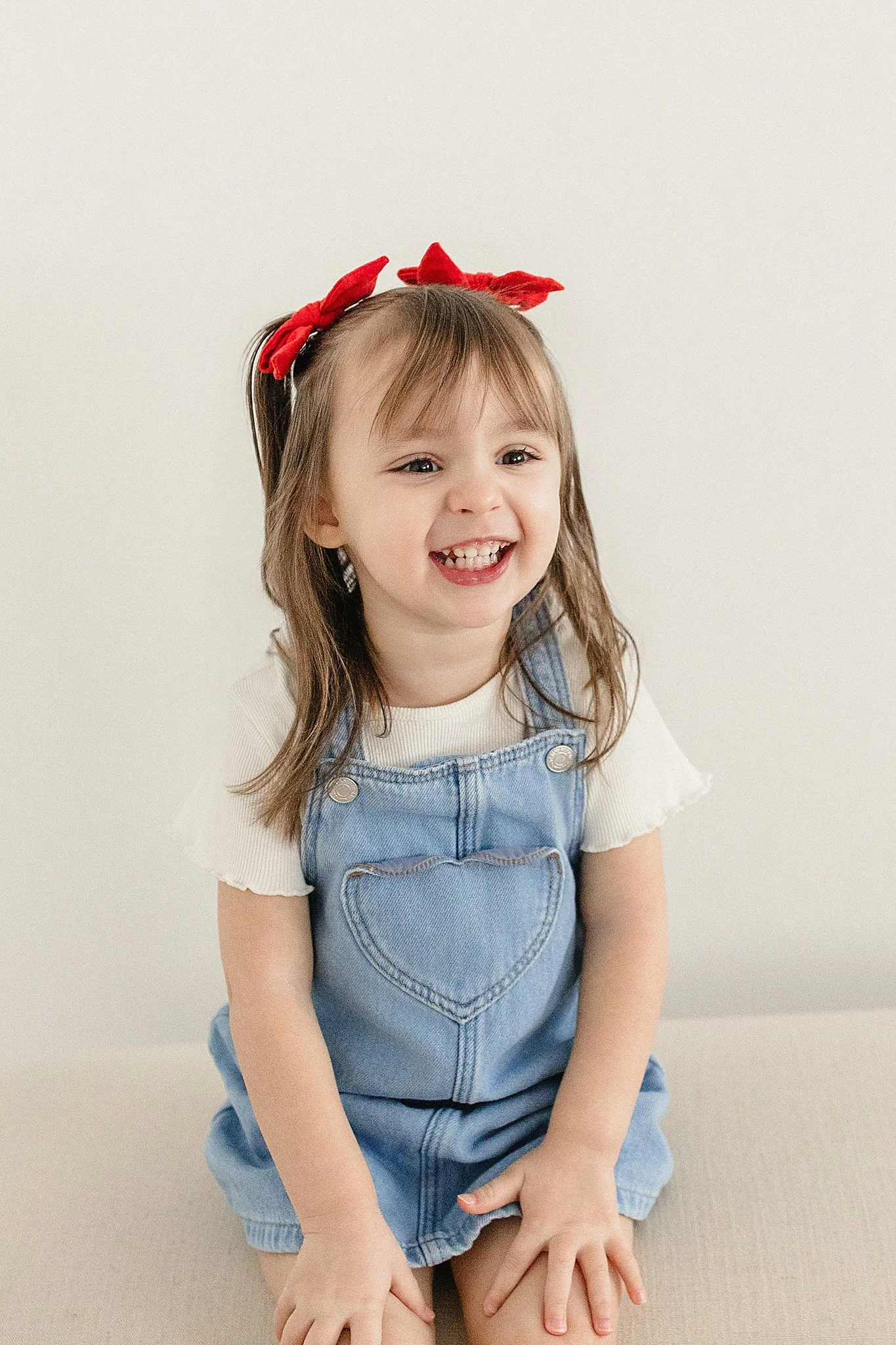 A young girl with brown hair, wearing a red bow headband, a white shirt, and denim overalls, smiling and sitting on a beige bench against a plain white wall for kids photos near me.