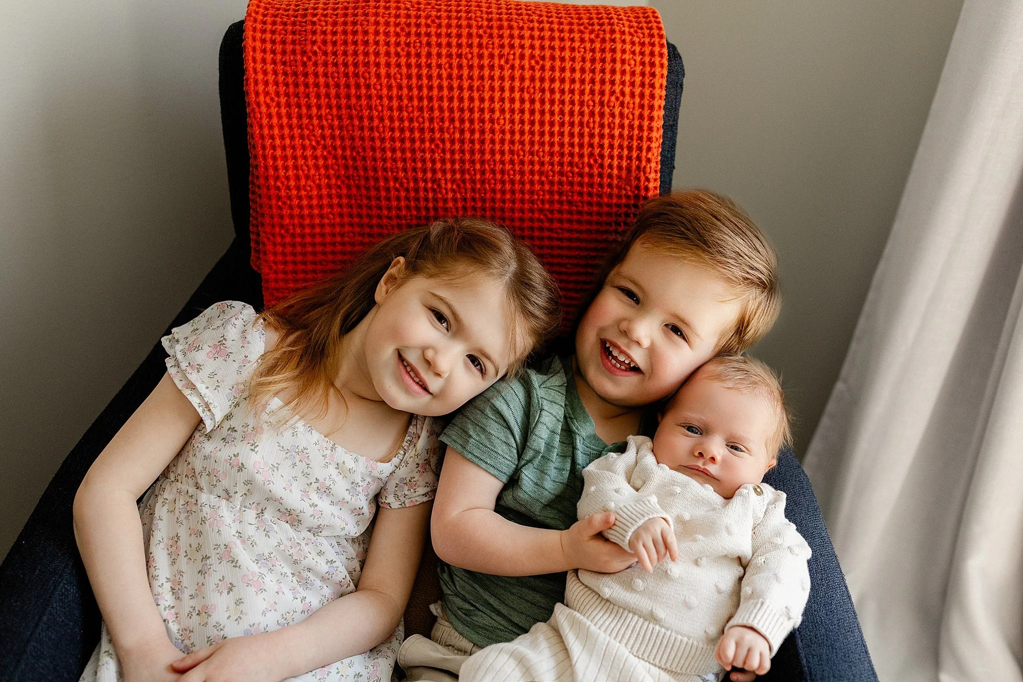 Four smiling children lying together on a chair, with a red blanket on the headrest. The children are diverse in appearance, with different hair colors and styles, and are wearing casual clothes for in home newborn photography near me.