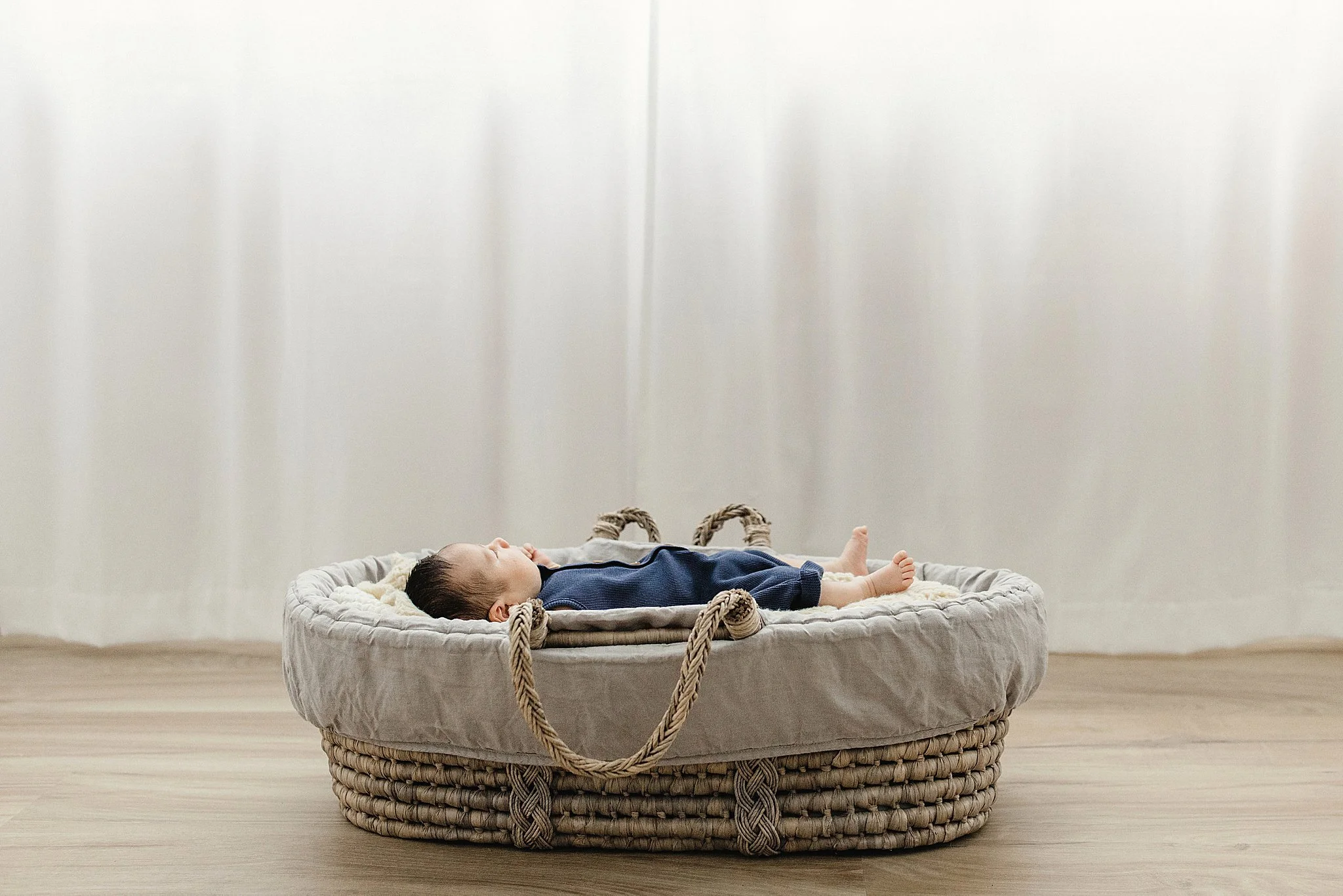 A baby lying on its back in a round, woven basket on a wooden floor, with a plain white wall in the background during a Naperville newborn photoshoot.