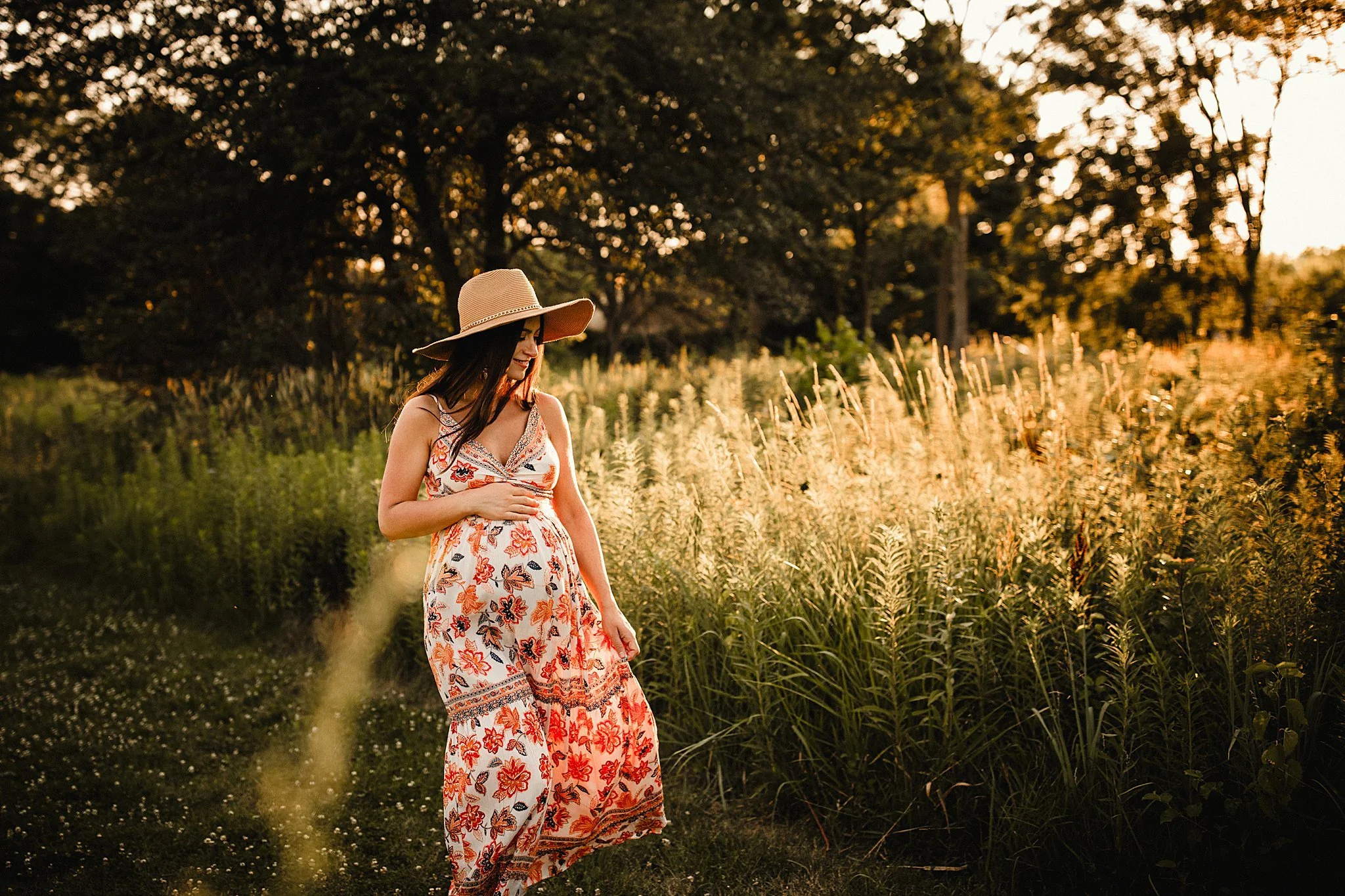 A woman wearing a floral dress and a large sun hat is walking through a grassy field during sunset. Trees are visible in the background during maternity photos near me.