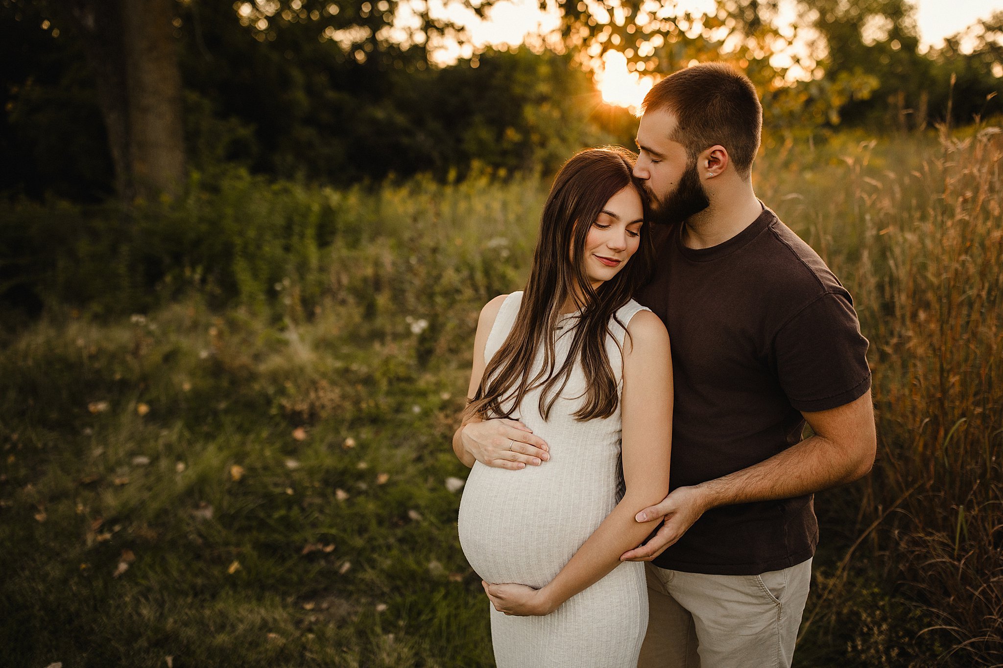 A pregnant woman and her partner stand outside at sunset, embracing each other in a grassy field with trees in the background for maternity photos near Naperville, IL
