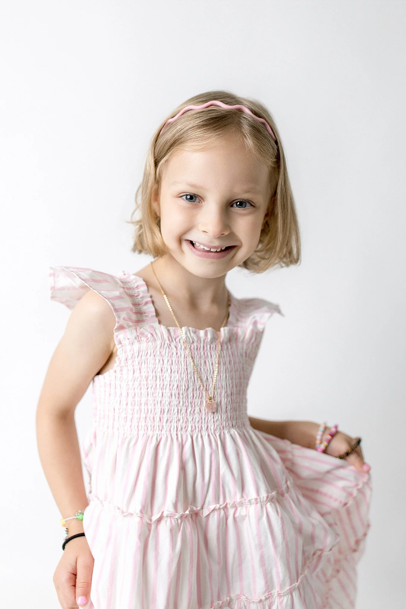 A young girl with blonde hair wearing a pink and white striped dress, smiling and looking at the camera, against a plain white background for the best kids photographer Naperville, IL.