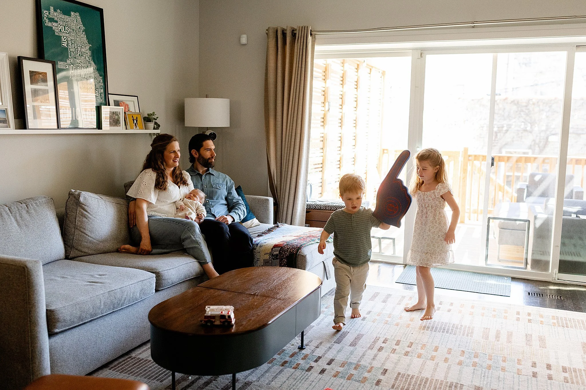 A family in a living room, with a woman, a man, and infants sitting on a sofa, and two children playing with a toy near the glass sliding door.