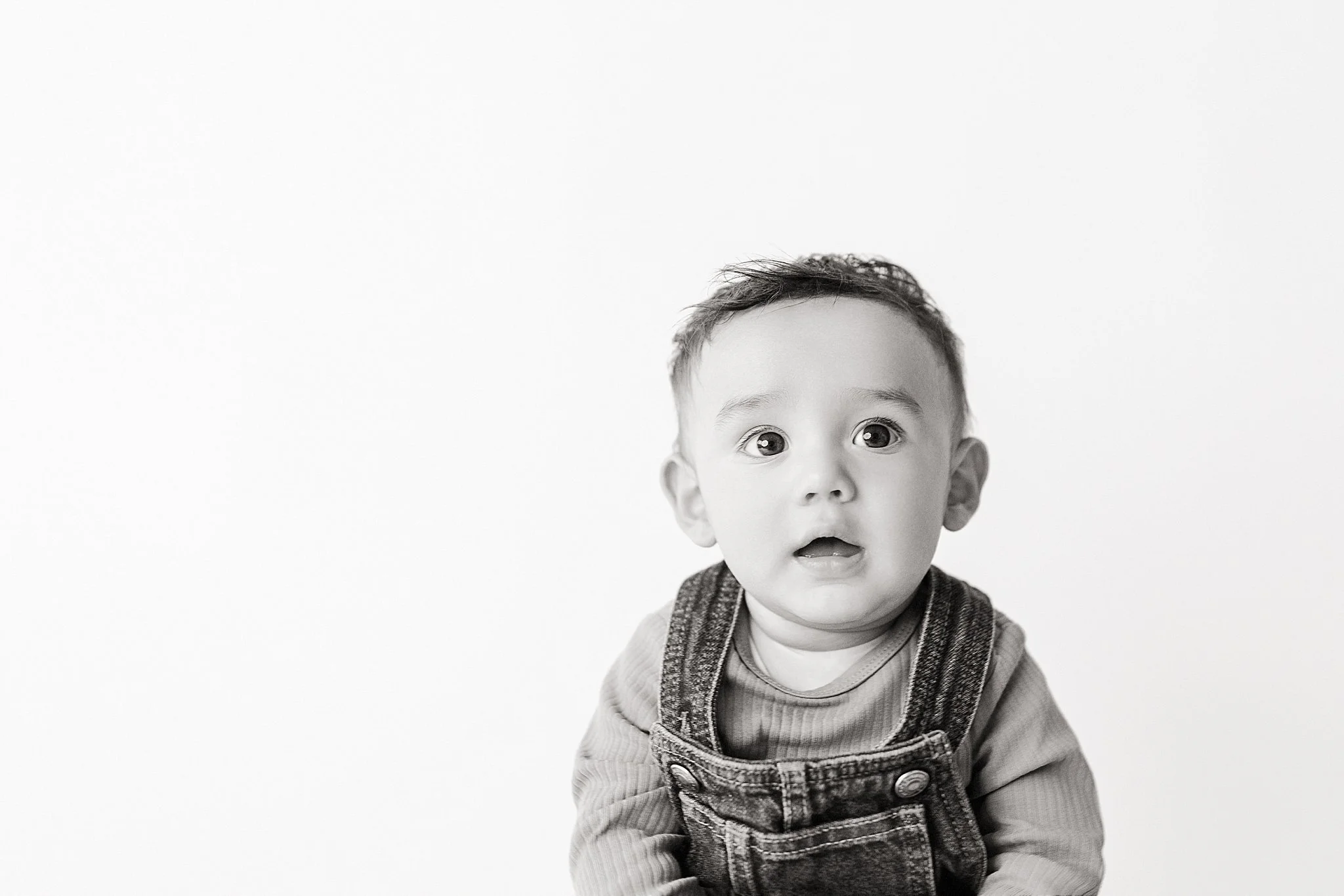 Black and white photo of a young child with a surprised or curious expression, wearing a denim overall and a long-sleeve shirt, against a plain background.