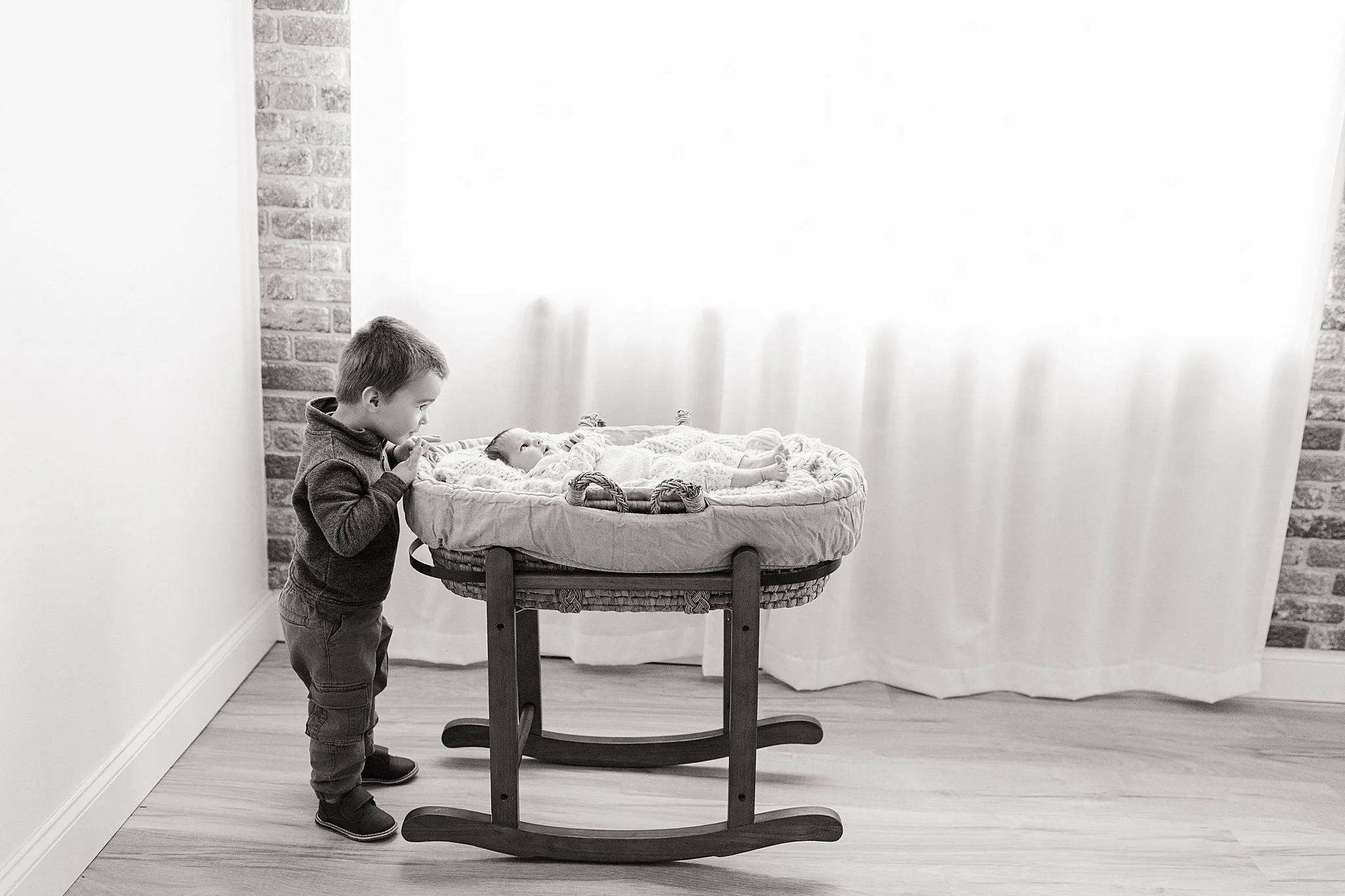 A young boy standing next to a bassinet with a baby inside, both looking at each other in a room with white walls and a brick accent wall during a studio lifestyle newborn photography session.