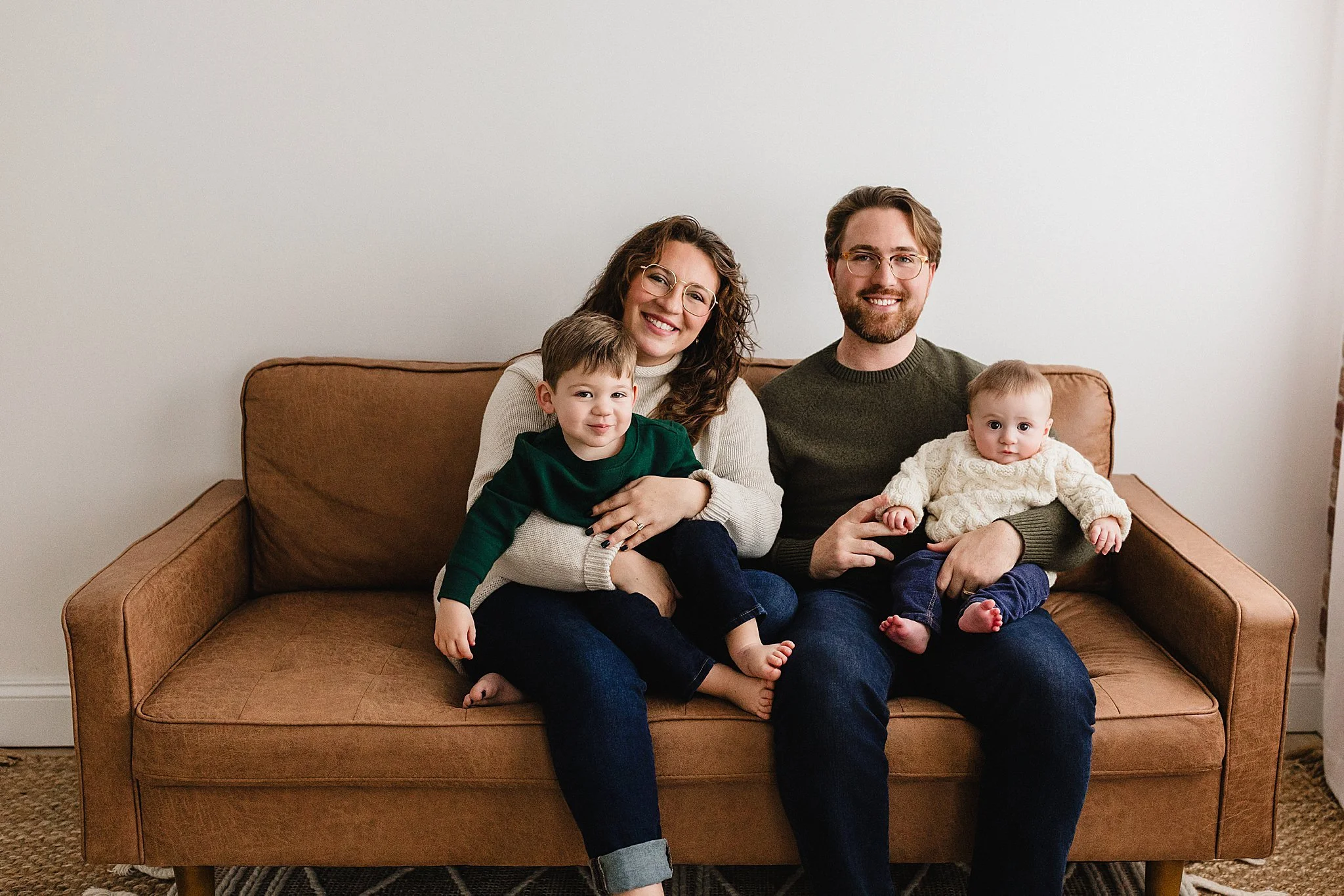 A smiling family of four sitting on a brown sofa in a living room with a plain white wall behind them during a Naperville studio family photoshoot.
