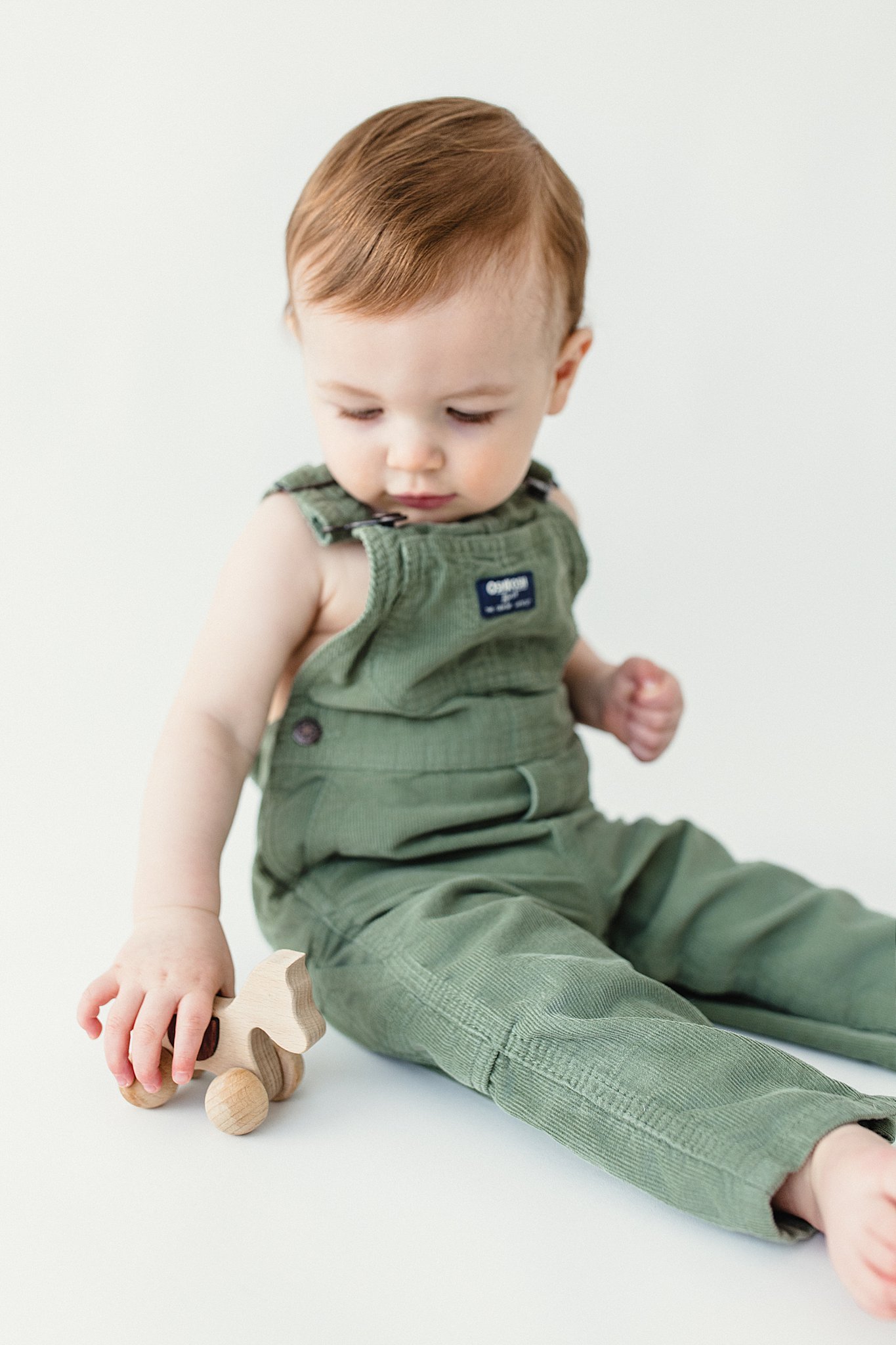 A young child with red hair wearing green overalls, sitting on a white surface and holding a wooden toy dog during an infant photoshoot in Naperville.