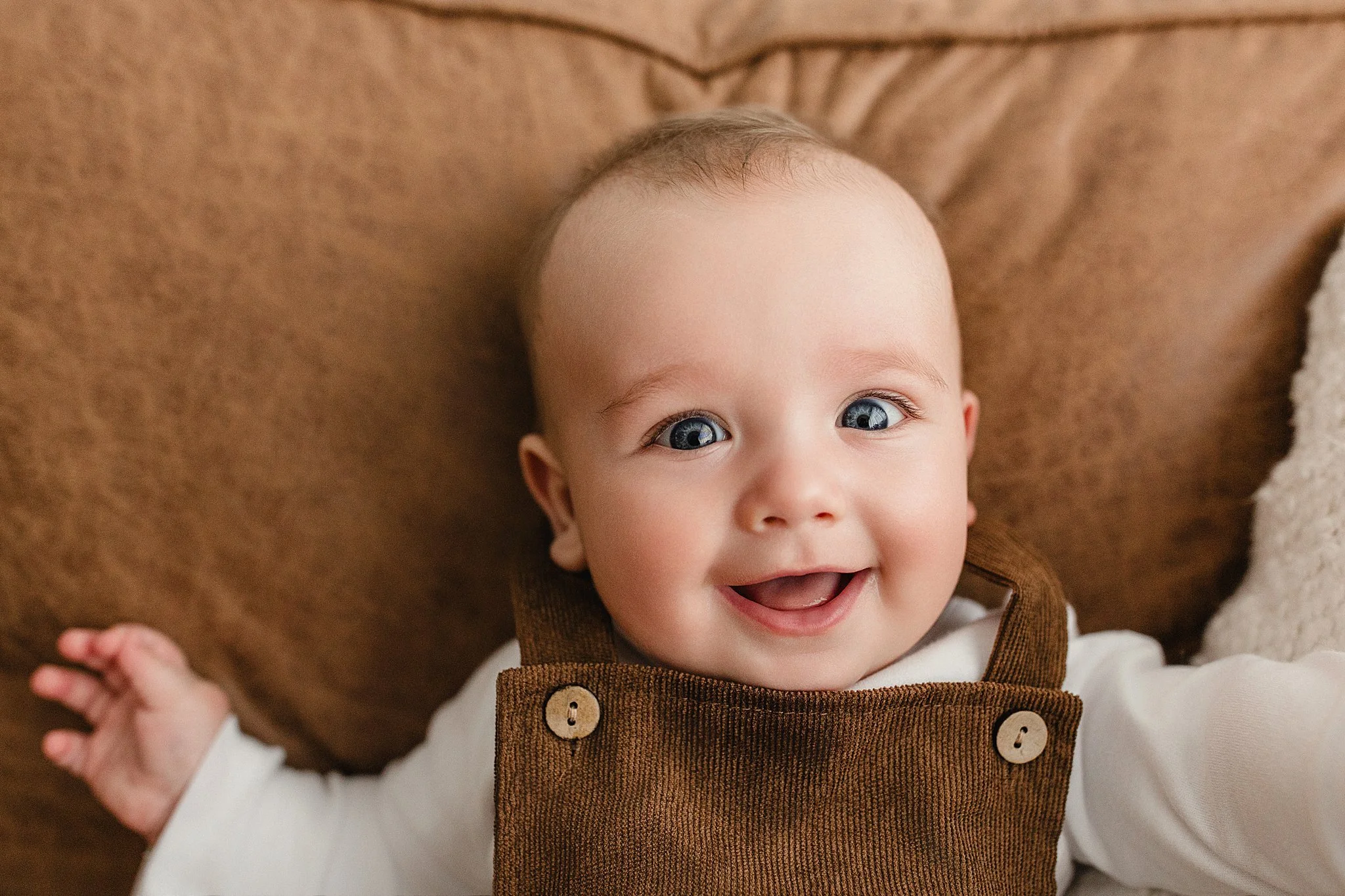 Smiling baby with blue eyes lying on a brown blanket, wearing a white shirt and brown overalls during a baby photo session in Naperville, IL.