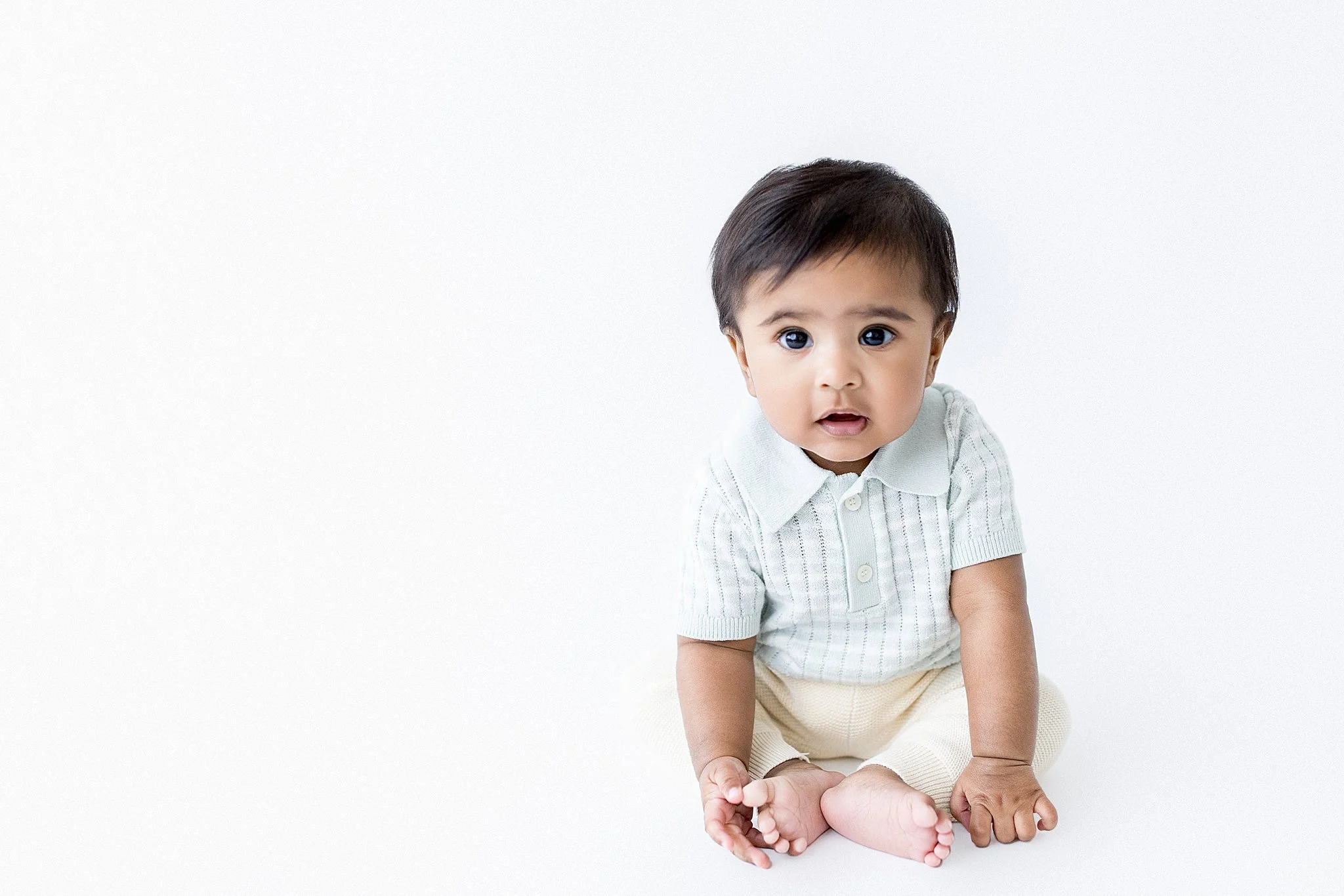 A baby with dark hair, wearing a light-colored shirt and pants, sitting on a white background, looking at the camera during a milestone photoshoot for a six month old baby in Naperville, IL.