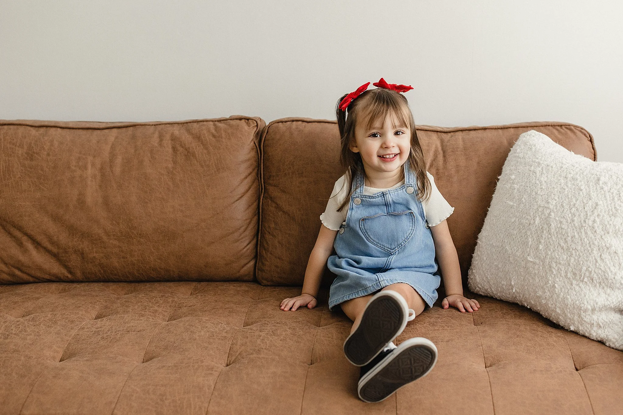 A young girl with a red bow in her hair, wearing a white shirt and denim overalls, sitting on a brown sofa next to a large white pillow, smiling at the camera for kids photography near me.