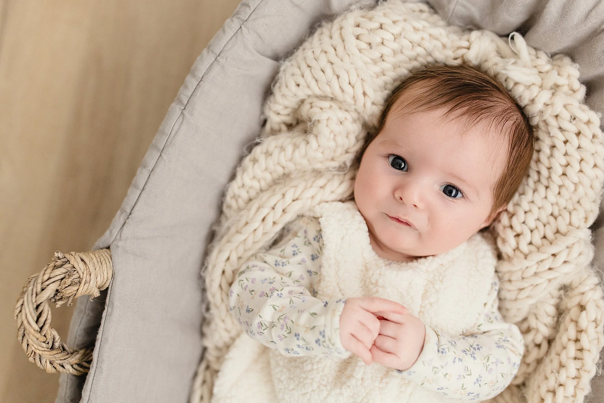 A baby with brown hair and blue eyes lying in a cozy, light-colored woven basket with a thick, cream-colored knitted blanket around the head, wearing a white long-sleeve shirt with a floral pattern and a cream-colored vest during newborn photos.