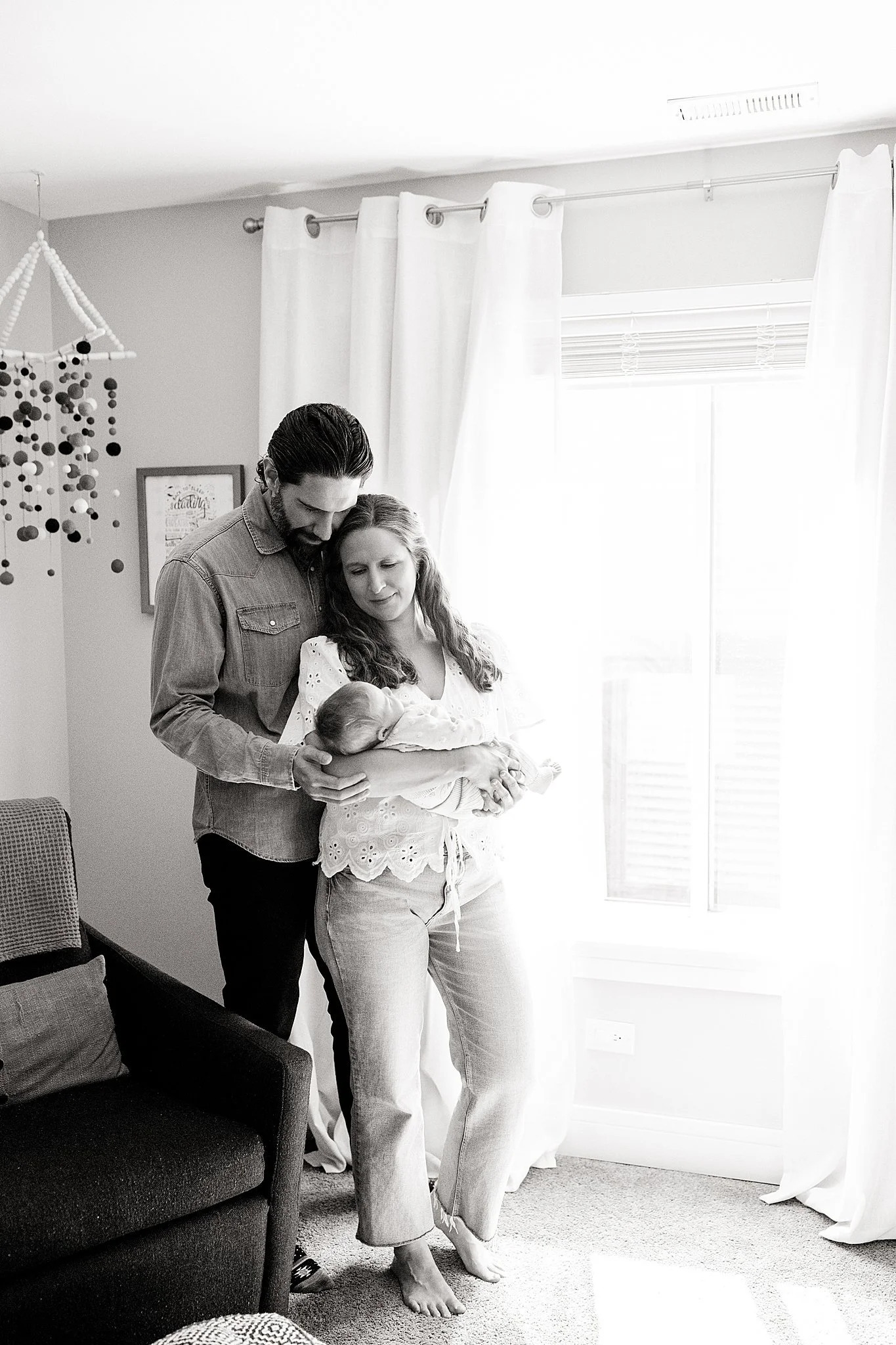 A family gathered in a living room, with a woman holding a newborn baby, and a man leaning in close, all looking at the infant. The room has curtains, a window, a framed picture on the wall, and a hanging mobile during a lifestyle shoot in Chicago.