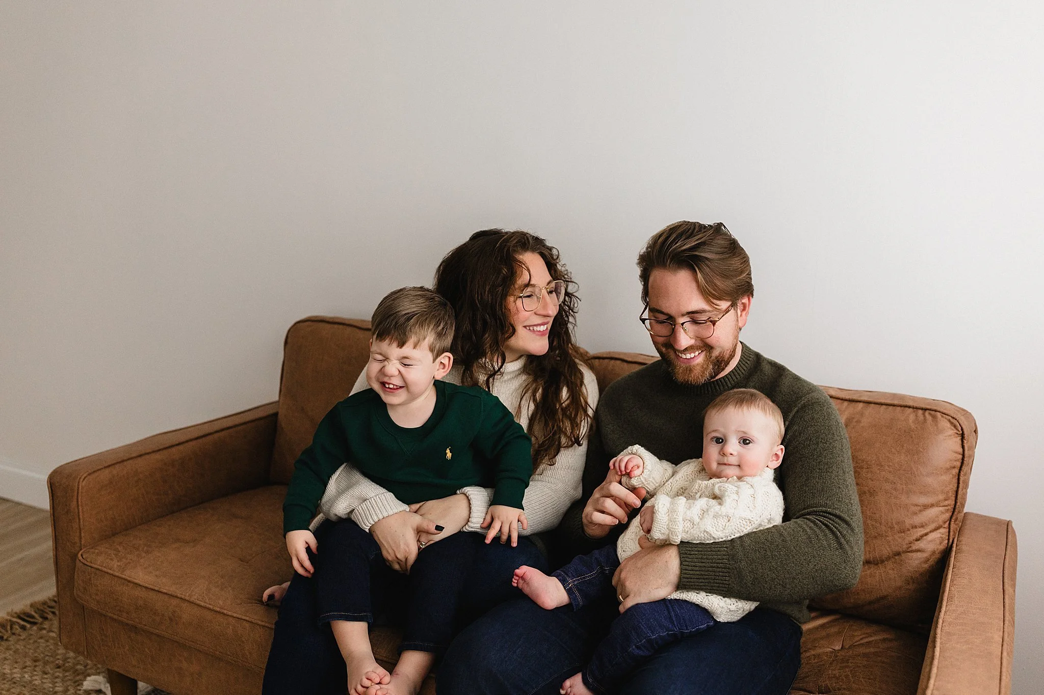 Family of four sitting on a brown sofa, smiling and enjoying time together. A woman with curly hair wearing glasses sits in the middle, with a young boy on her lap laughing. Next to her, a man with glasses holds a baby girl during family photos.