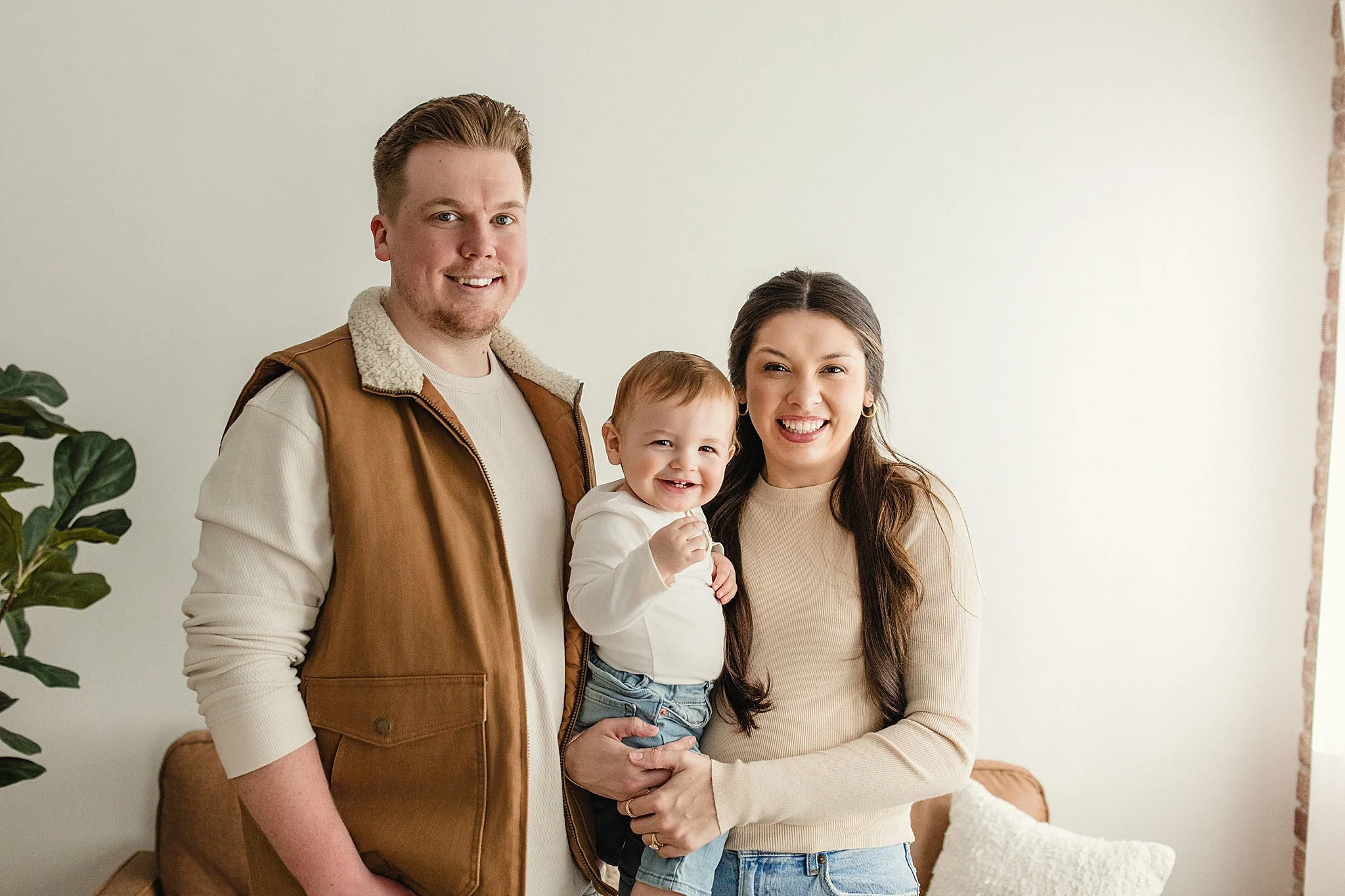 A family of three standing indoors smiling at the camera, with a man on the left, woman on the right holding a baby, a white wall in the background, and a plant on the left side.