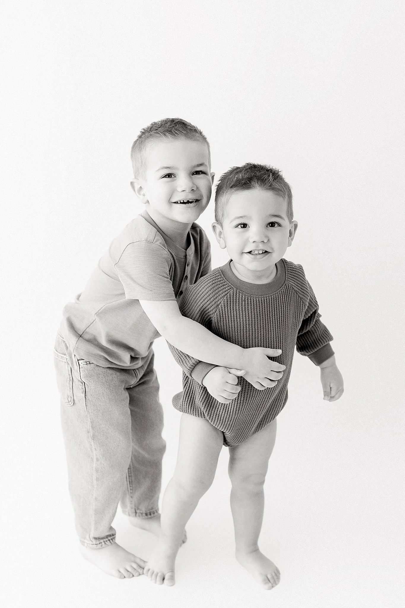 Two young boys smiling, hugging each other, and standing barefoot against a plain background black and white kids photography with Ally and B Photography.