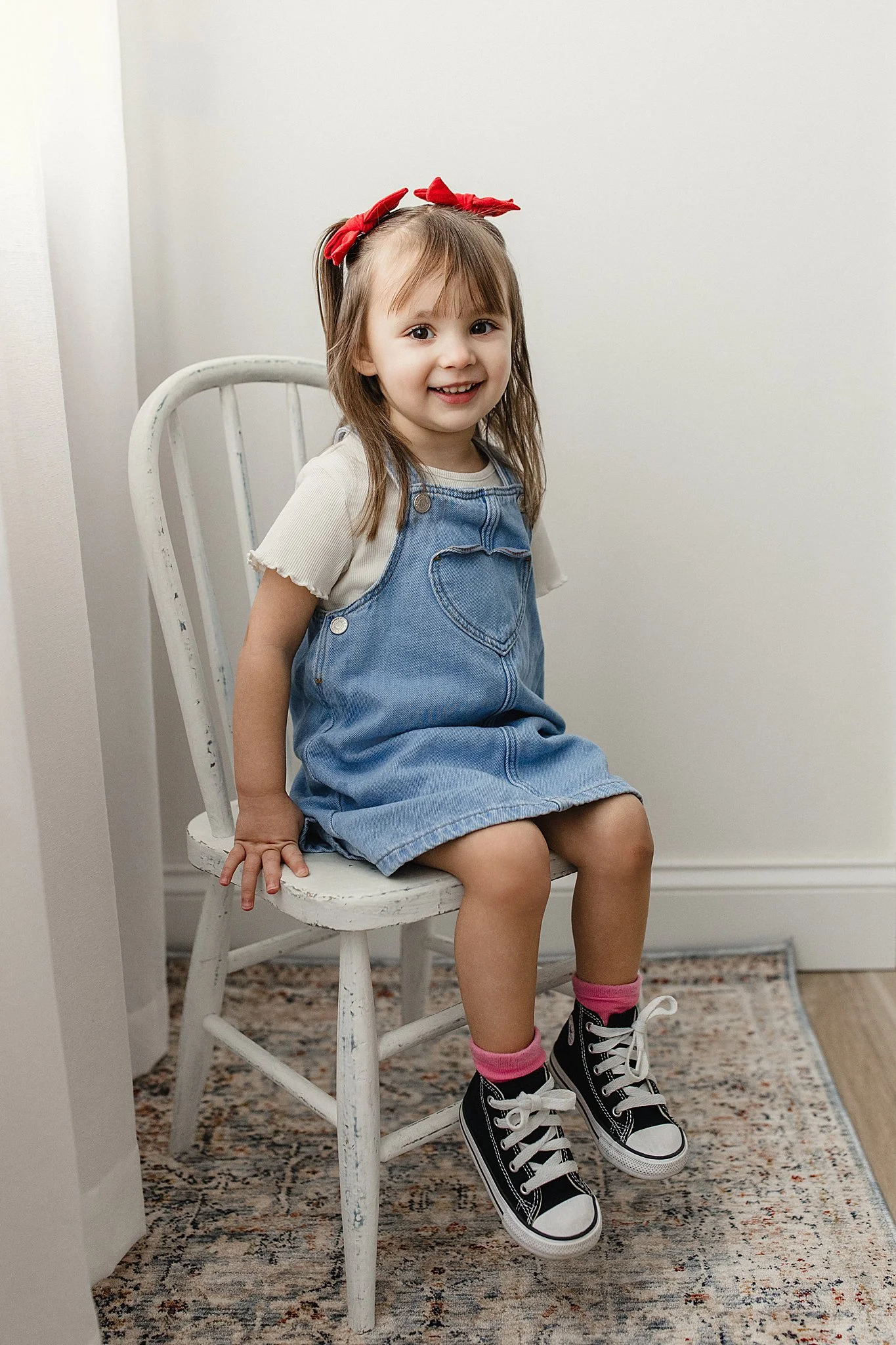 A young girl sitting on a white wooden chair with a smile, wearing a denim overall dress, beige t-shirt, pink and black sneakers, pink socks, and red hair ribbons during a children's photoshoot near Naperville, IL.