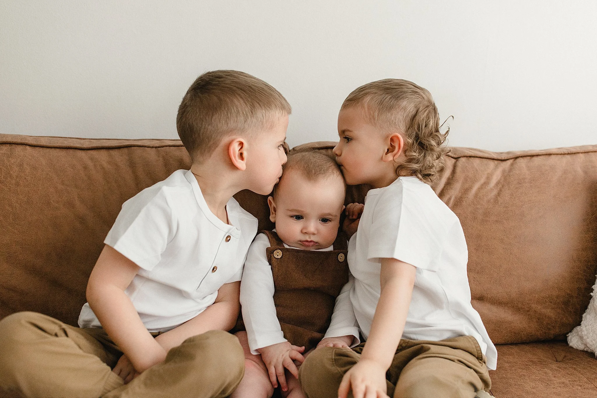 Three young children sitting on a brown couch, with two older boys kissing a baby on the head during a lifestyle family photoshoot with Ally and B Photography in Naperville, IL.