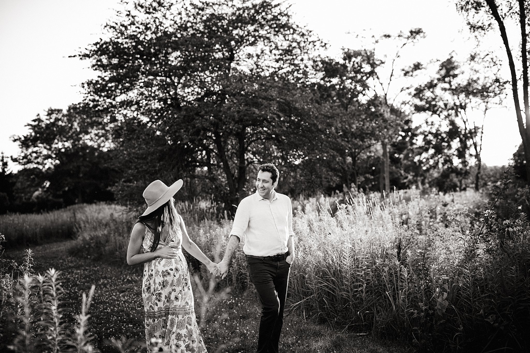 A black and white photo of a couple walking hand in hand outdoors, surrounded by trees and tall grass, with the woman wearing a wide-brimmed hat and a floral dress, and the man smiling for Naperville maternity photos.