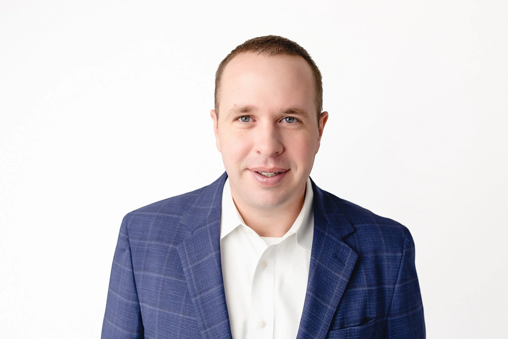 Headshot of a man with short brown hair, blue eyes, wearing a dark blue blazer and white collared shirt, smiling gently against a plain white background with a Naperville headshot photographer.