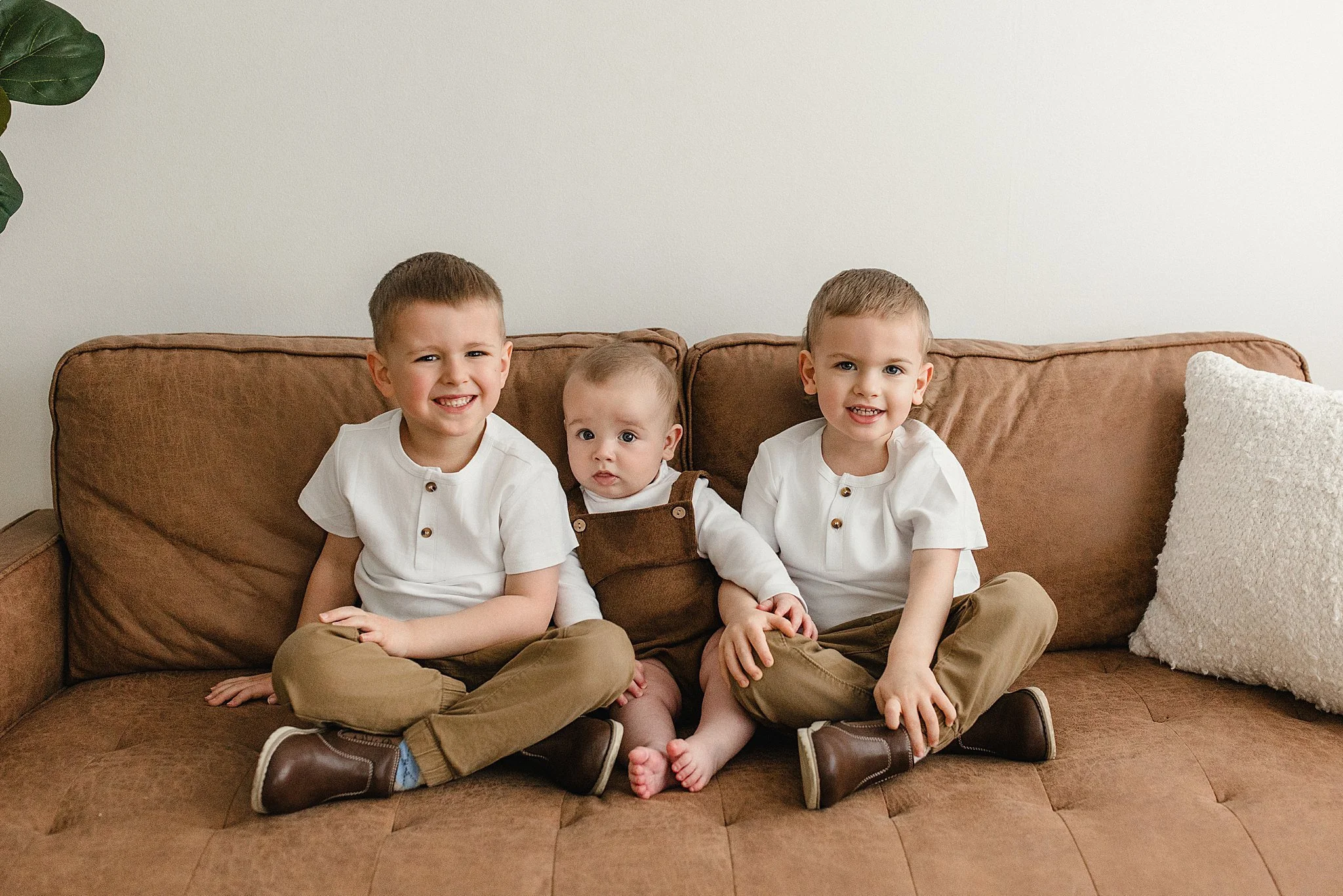 Three young boys sitting on a brown couch against a plain wall. They are dressed in white tops, brown pants, and similar brown shoes. The youngest in the middle is wearing brown overalls. They are all smiling during family photos in Naperville.